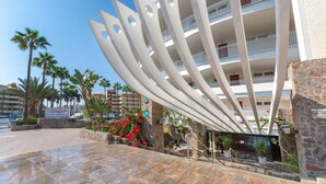 Un hotel de varios pisos con balcones, fachada de piedra y un área de entrada pavimentada con palmeras y flores.