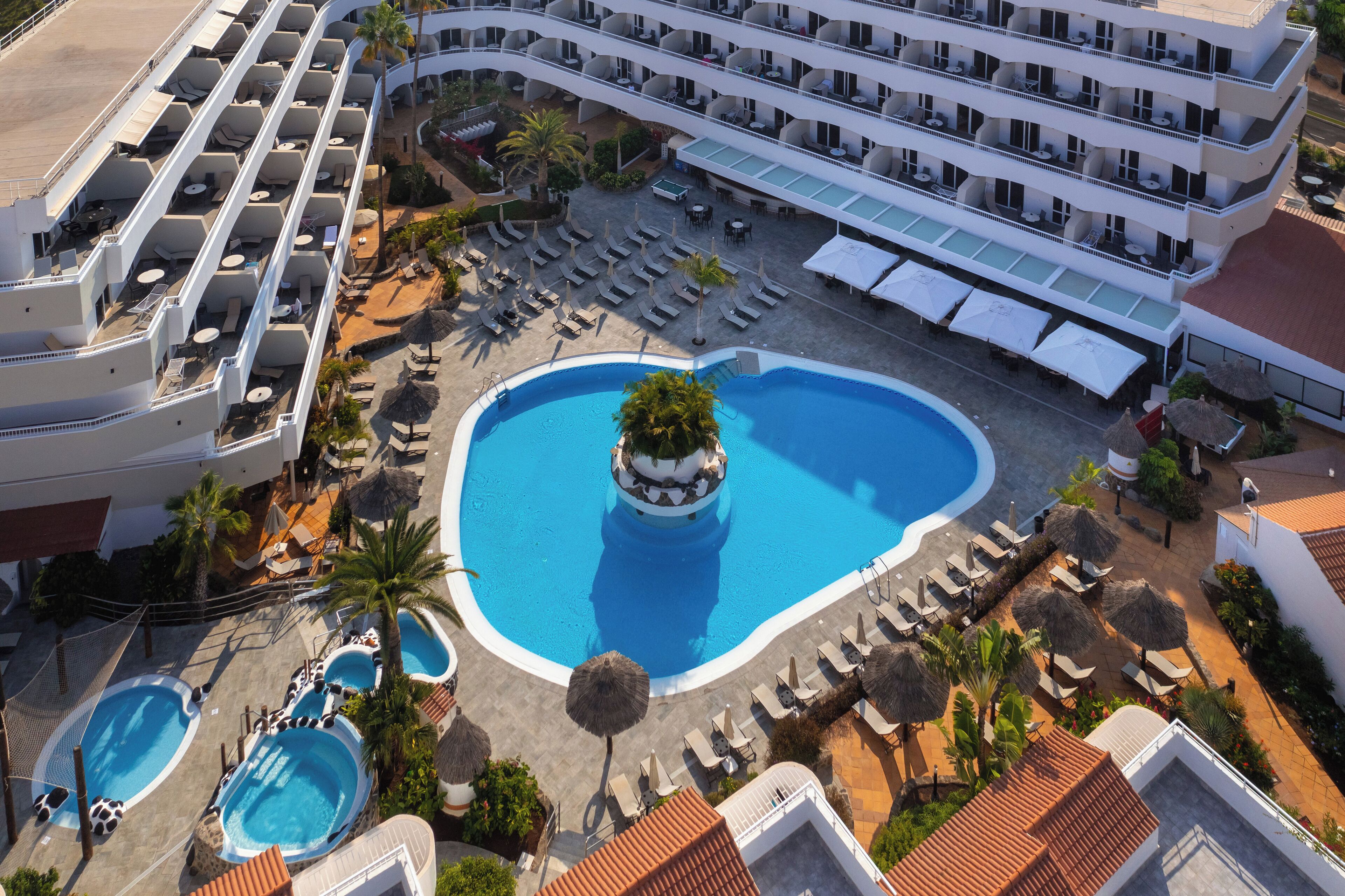 A hotel with multiple swimming pools, sun loungers, and a central pool with a decorative island.