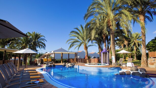 Una piscina con sillas de descanso y sombrillas, rodeada de palmeras.
