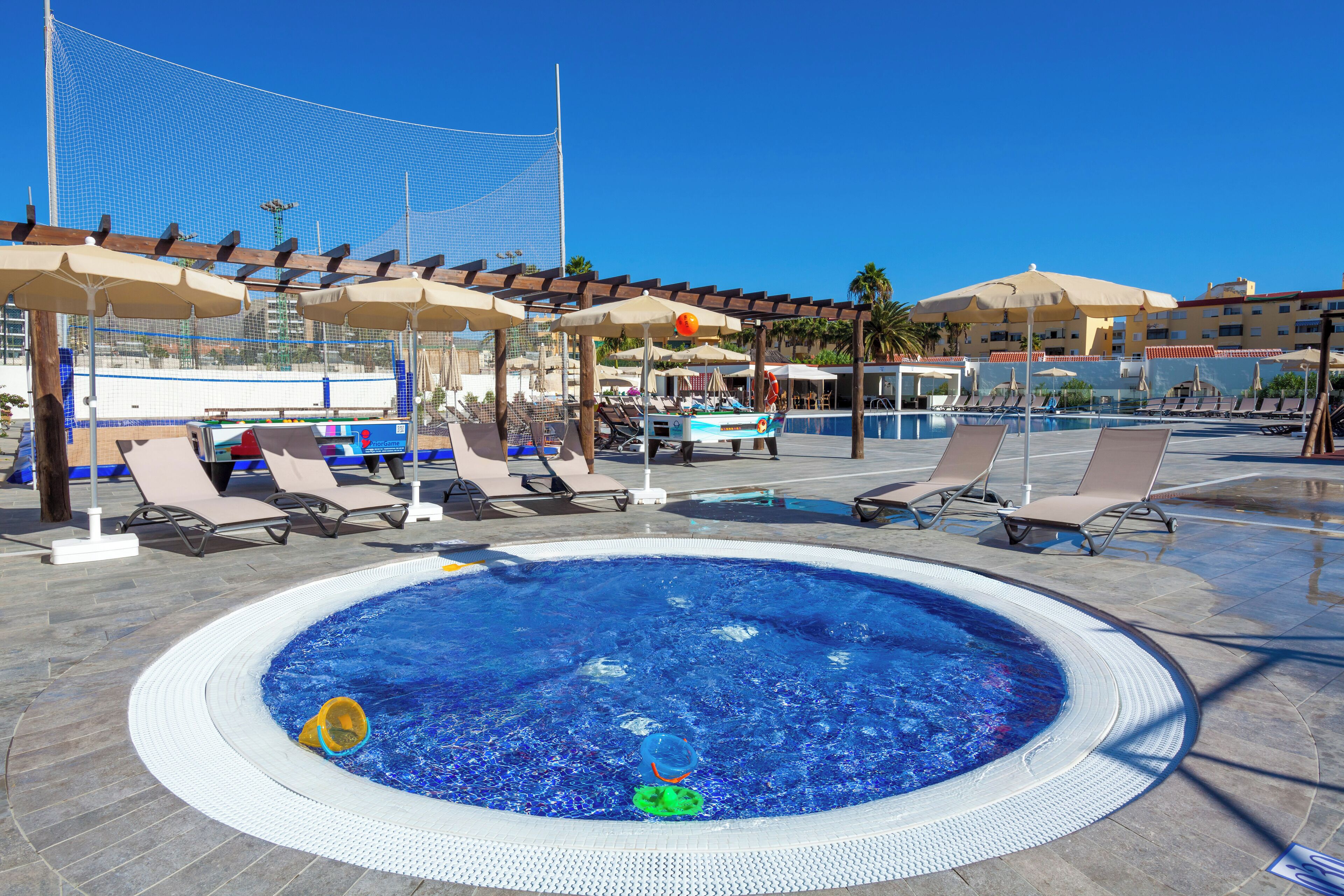 A swimming pool with lounge chairs and umbrellas, surrounded by a netted area and palm trees.