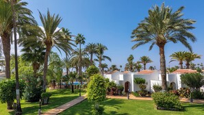 Un jardín cuidado con palmeras, un sendero pavimentado y edificios blancos con arcos.