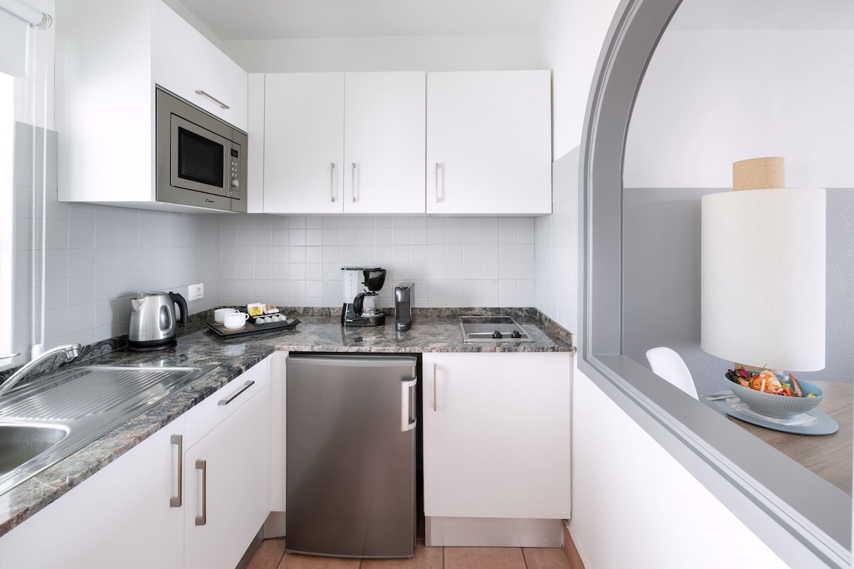 Una cocina compacta con armarios blancos, un refrigerador de acero inoxidable, un microondas, una cafetera y un lavamanos.