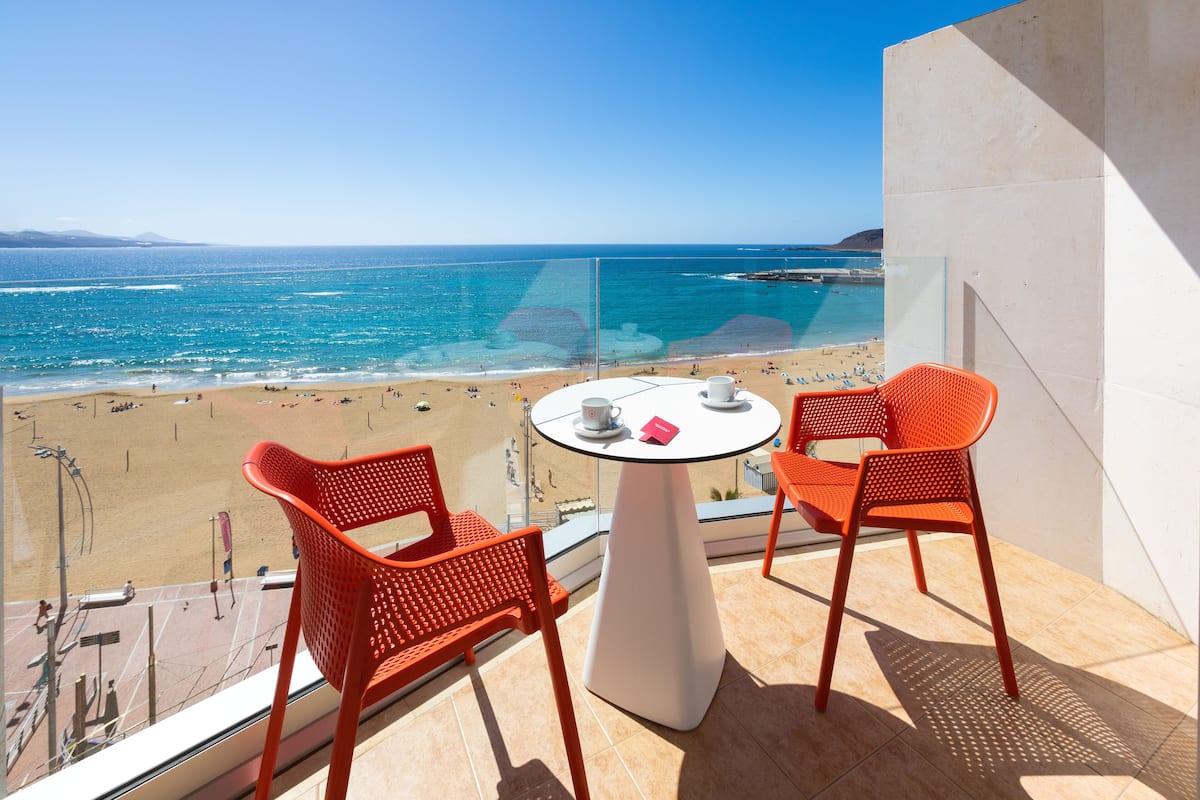 A balcony with two orange chairs and a small table, overlooking a beach with clear blue water and a distant shoreline.