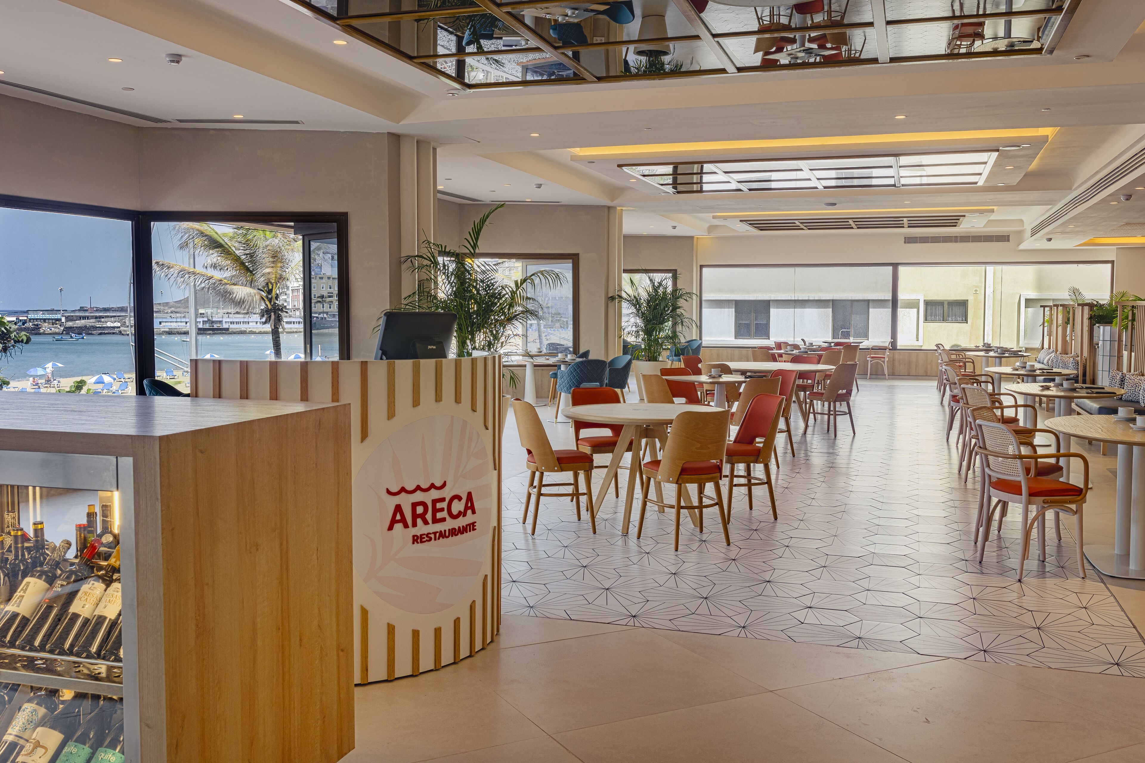 En moderne restaurant med havudsigt, med en træreception, flere spiseområder med stole og borde, samt store vinduer.