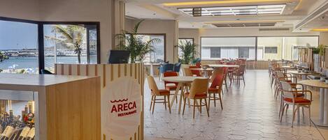 En moderne restaurant med havudsigt, med en træreception, flere spiseområder med stole og borde, samt store vinduer.
