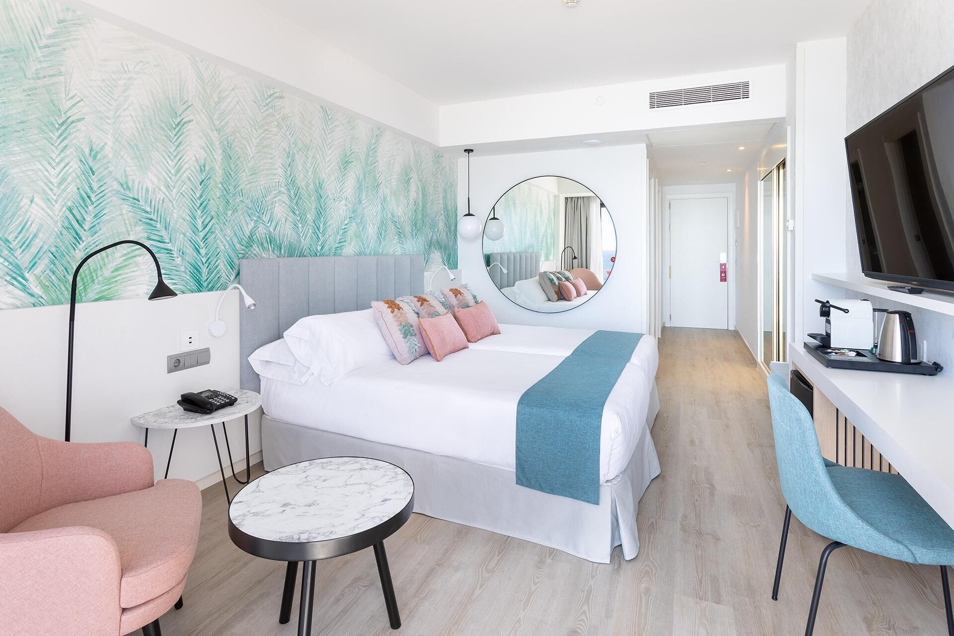 A modern hotel room with a bed, a round mirror, a desk with a TV, a chair, and a small table.