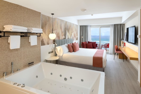 A hotel room with a large bathtub, a bed with red and white bedding, a desk with a chair, and a view of the ocean.