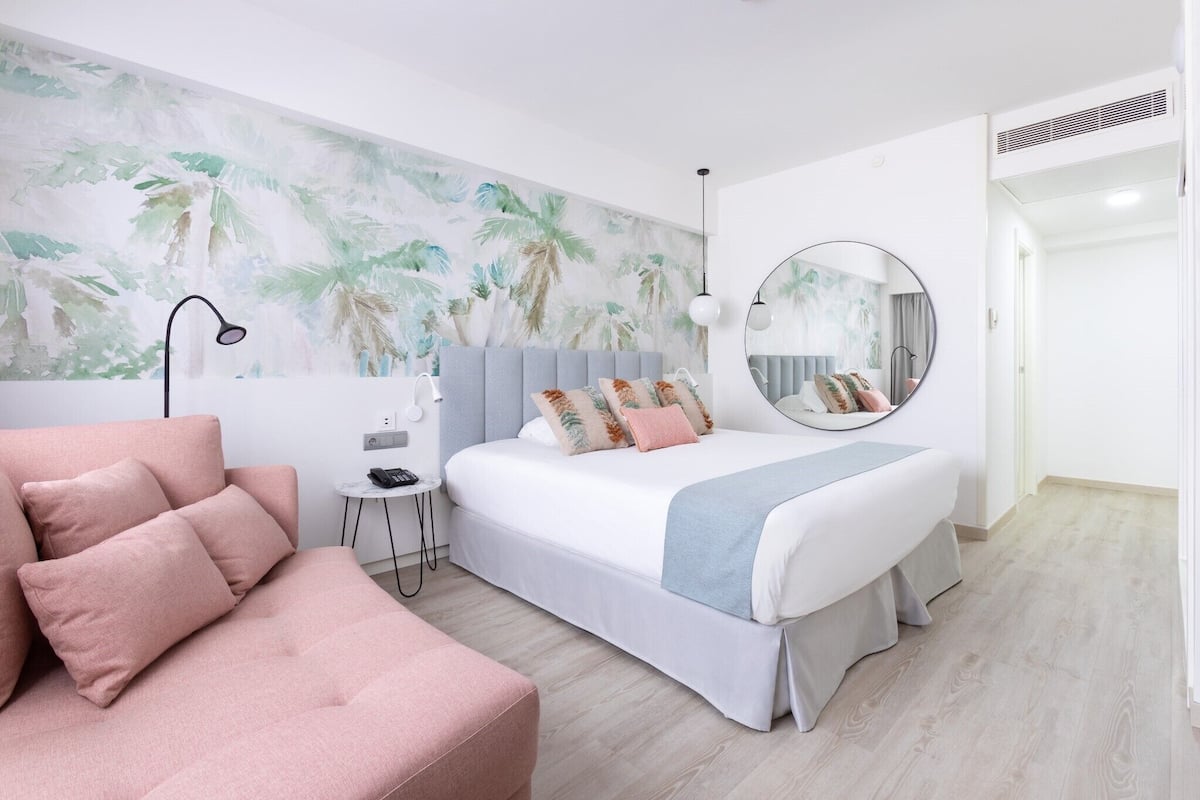 A modern bedroom with a bed, a pink sofa, a small side table, a round mirror, and a wall with a tropical mural.