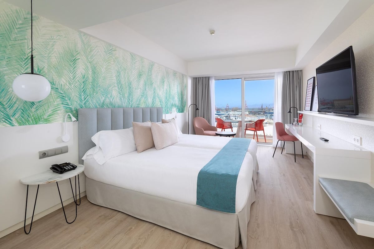 A hotel room with a large bed, a desk, a chair, a TV, and a view of the city.