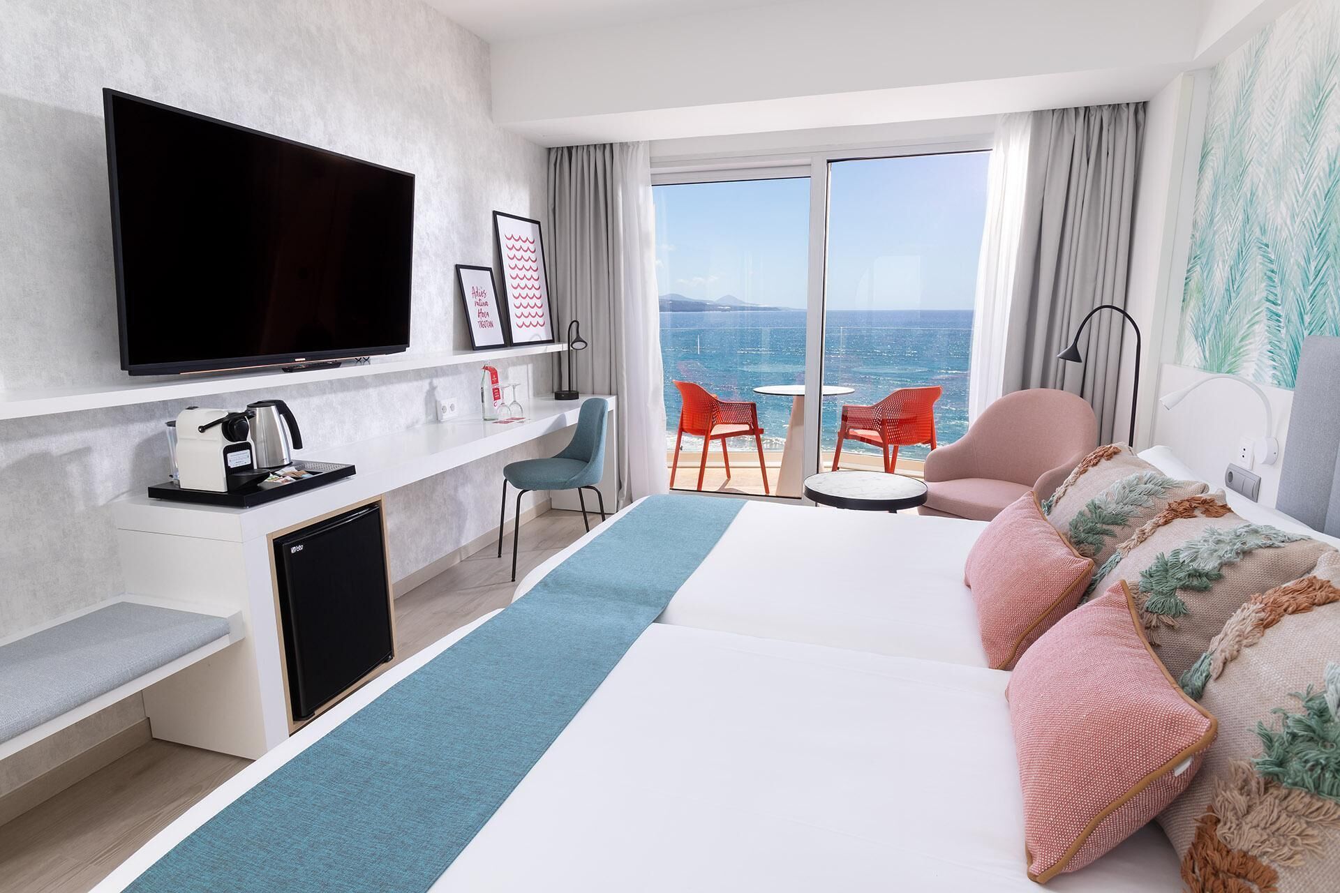 A hotel room with a large bed, a desk with a TV, a coffee machine, and a view of the sea through large windows.