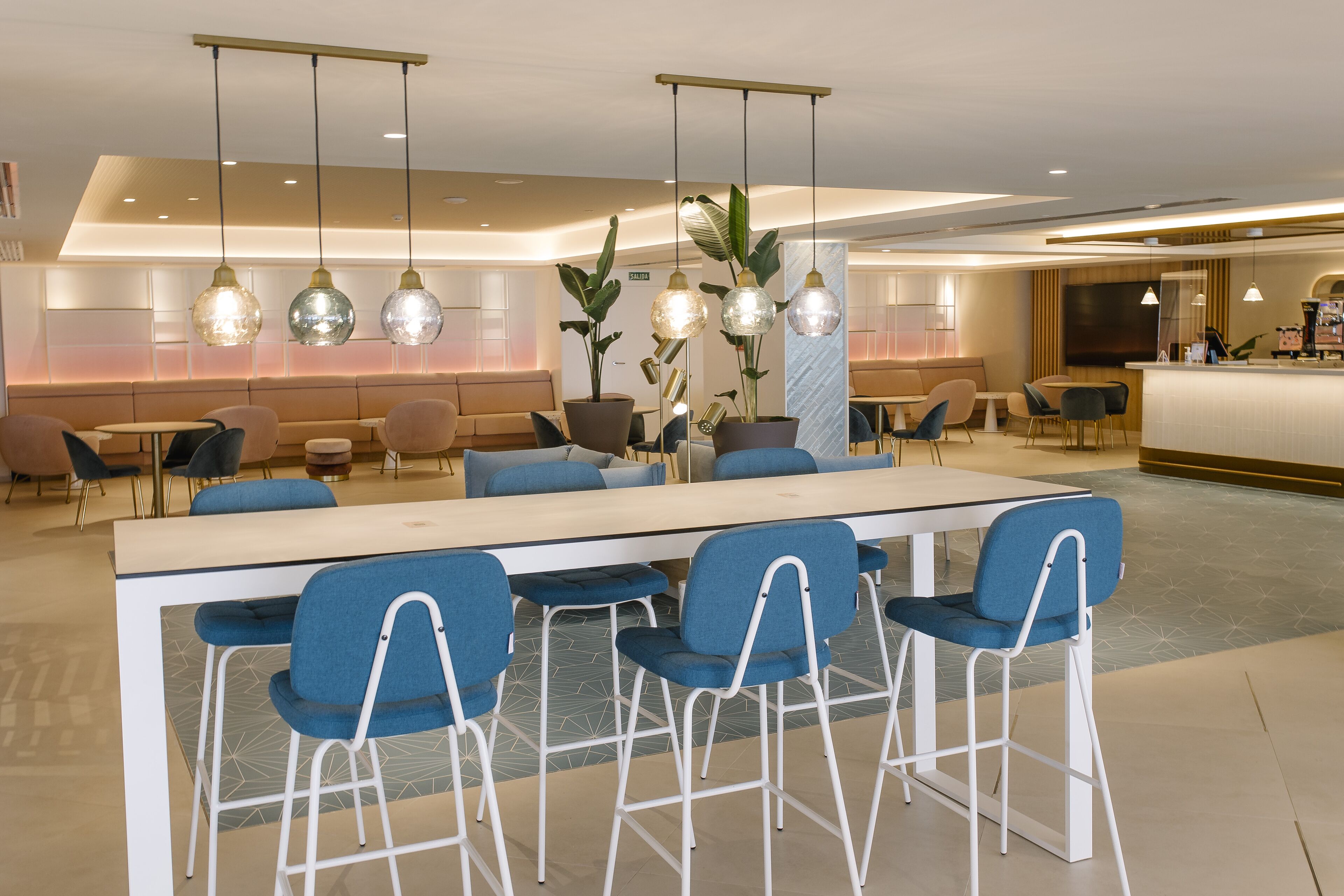 A modern hotel lobby with a long reception desk, blue bar stools, and hanging pendant lights.