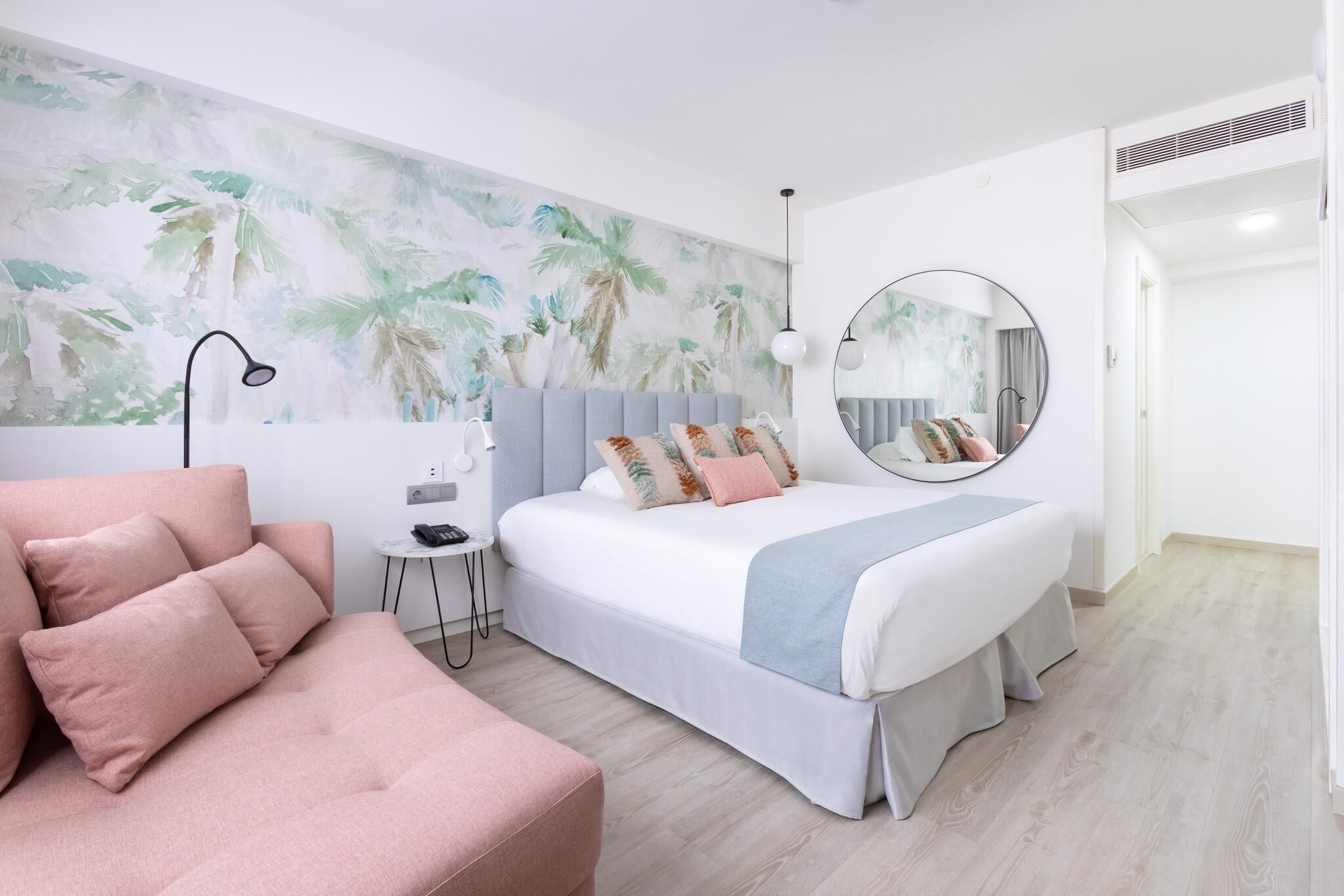 A modern bedroom with a bed, a pink sofa, a small side table, a round mirror, and a wall with a tropical mural.