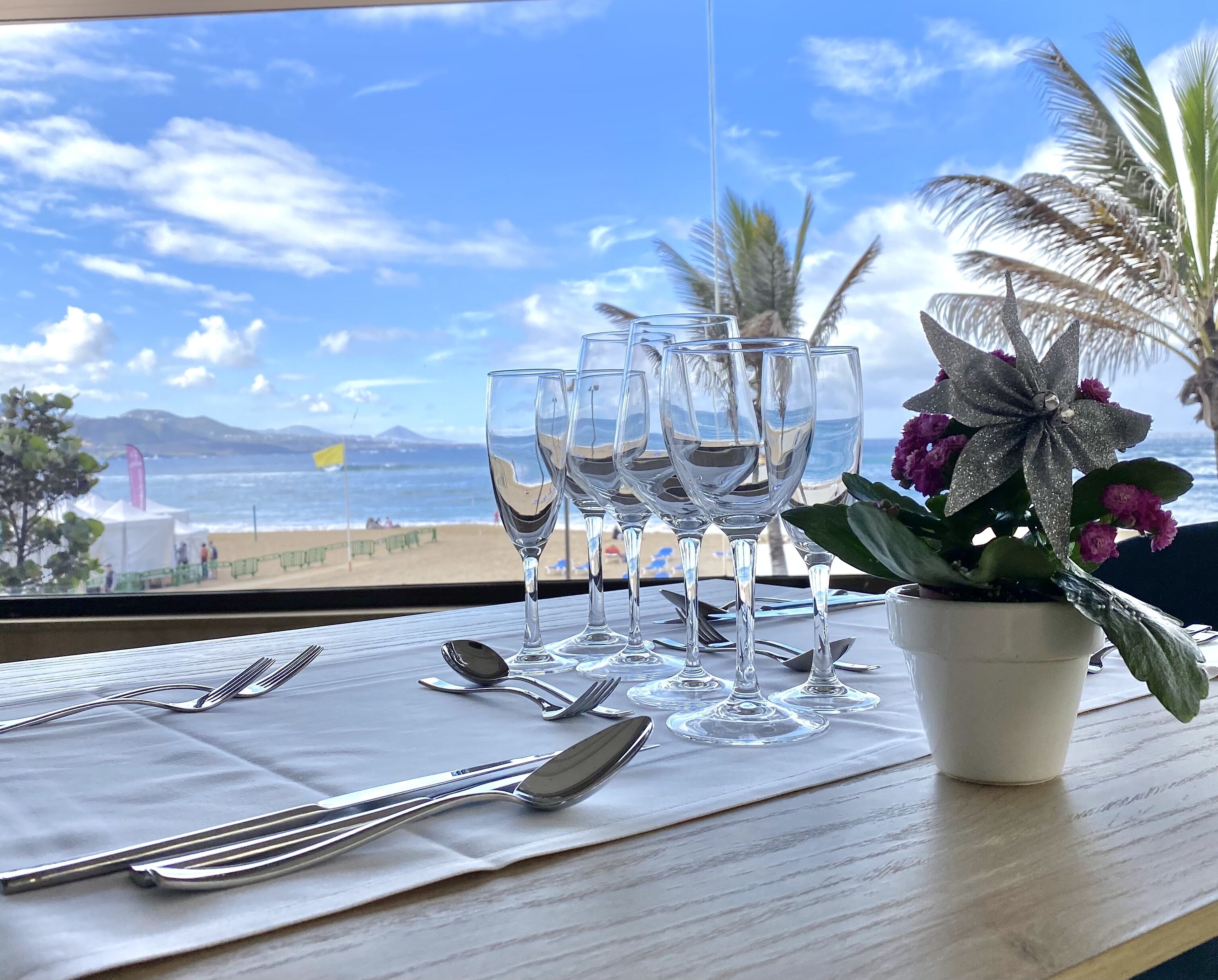 A table set with wine glasses, cutlery, and a flower arrangement, with a view of the beach and ocean.