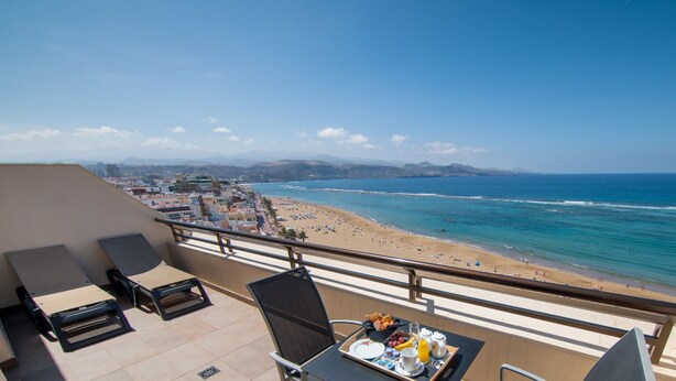 A balcony with a table set for breakfast, overlooking a beach with a clear view of the ocean and distant mountains.