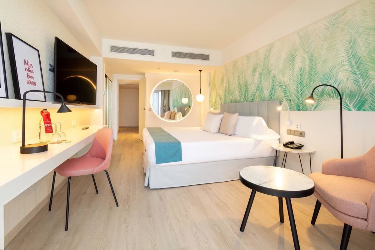 A modern hotel room with a bed, a desk, a chair, a round mirror, and a small table.