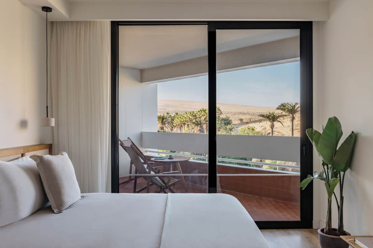 Een hotelkamer met een bed, een stoel, een tafel en uitzicht op een woestijnlandschap door een schuifdeur.