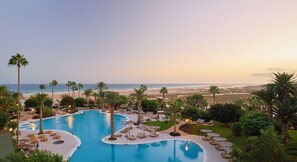 Zwembad van een resort met een golvend ontwerp, omringd door palmbomen en ligbedden, met een strand en uitzicht op de oceaan op de achtergrond.