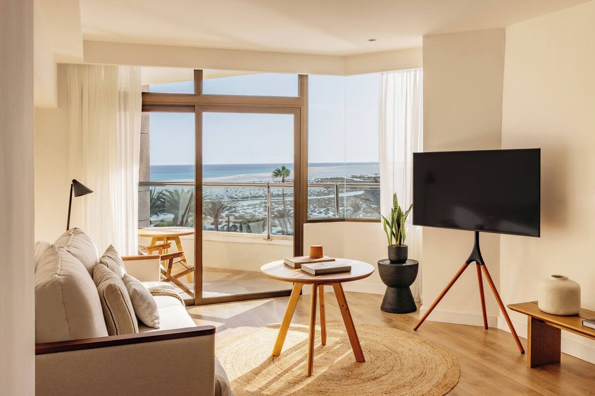 A modern living room with a large TV, a sofa, a small table, and a view of the beach.