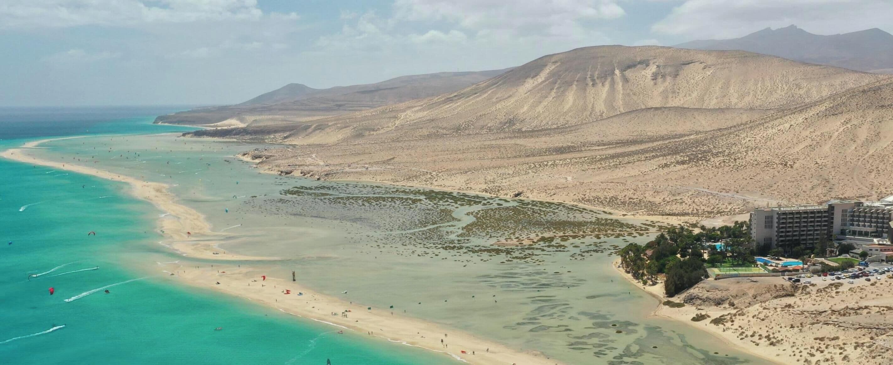Un paysage côtier avec une plage, des eaux turquoise et limpides, et une chaîne de montagnes en arrière-plan.