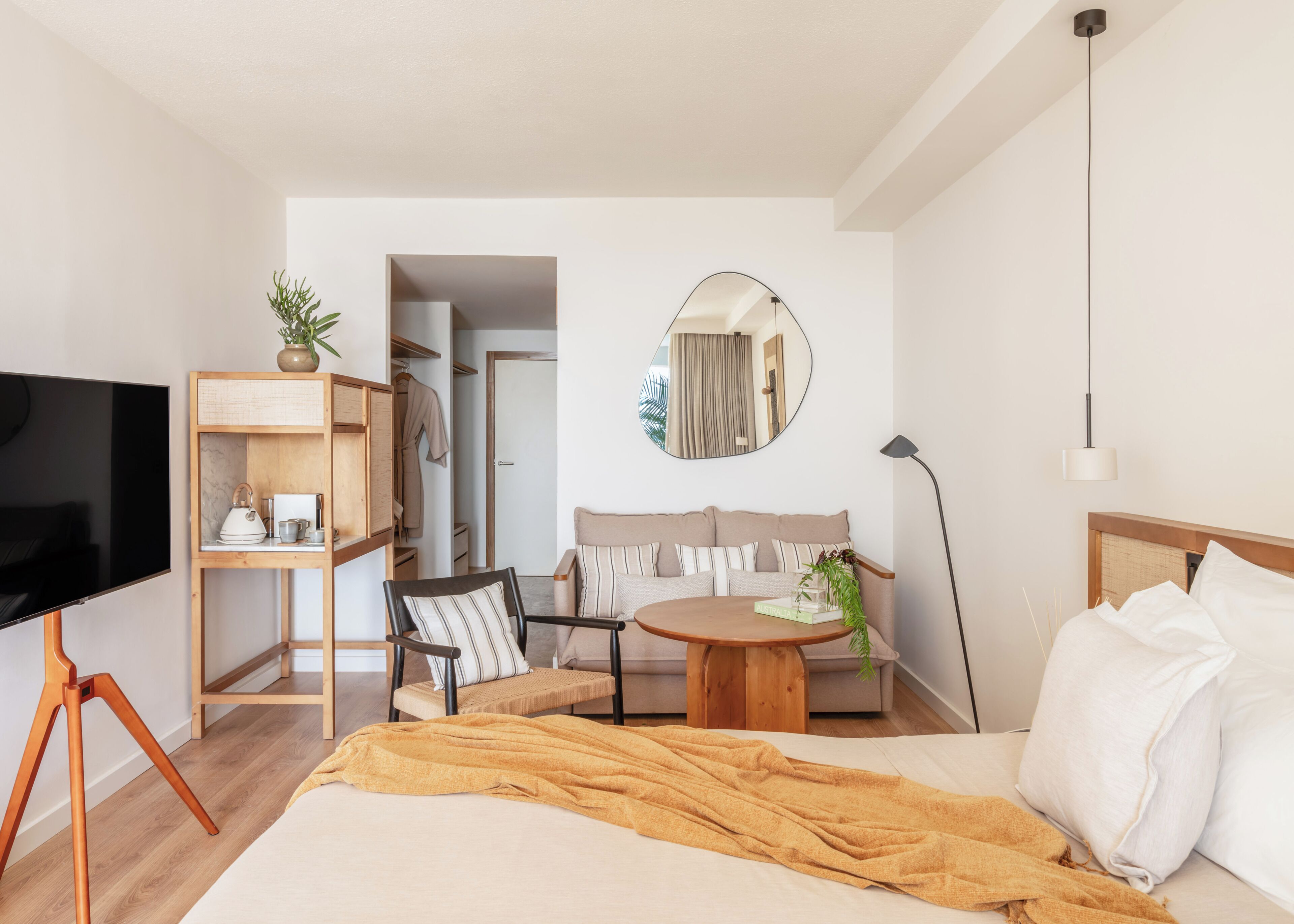 Una habitación de hotel moderna con cama, televisión, escritorio, silla, sofá, mesa redonda, espejo y planta.