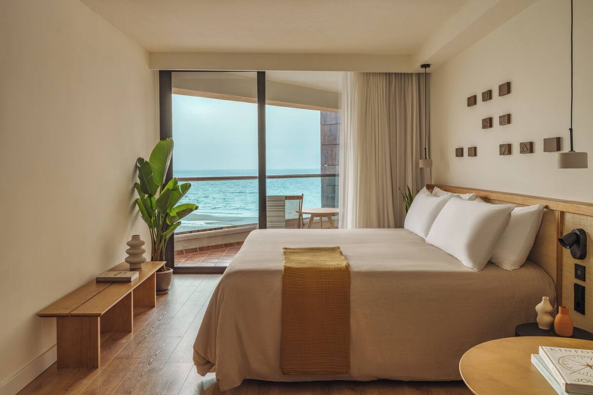A hotel room with a large bed, a wooden bench, a small table, and a view of the ocean.