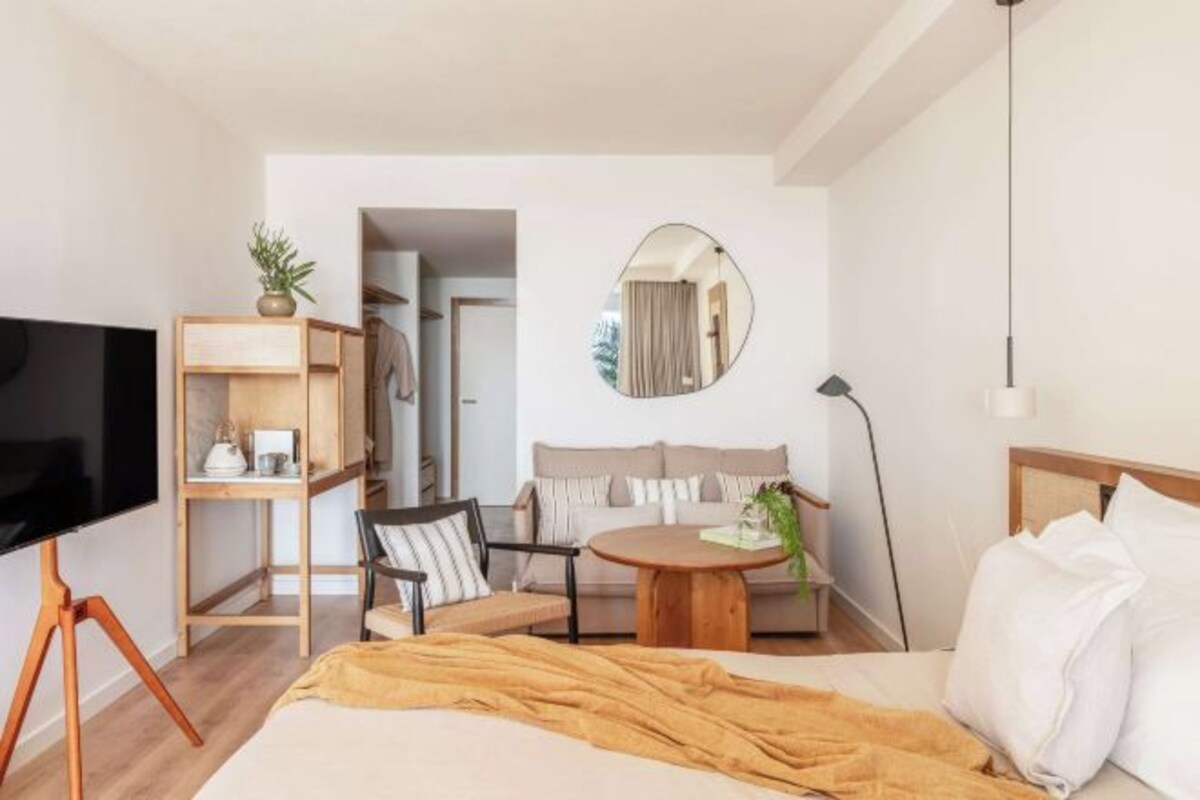 Een hotelkamer met een bed, een televisie op een standaard, een ronde salontafel, een bank, een stoel, een spiegel en een lamp.