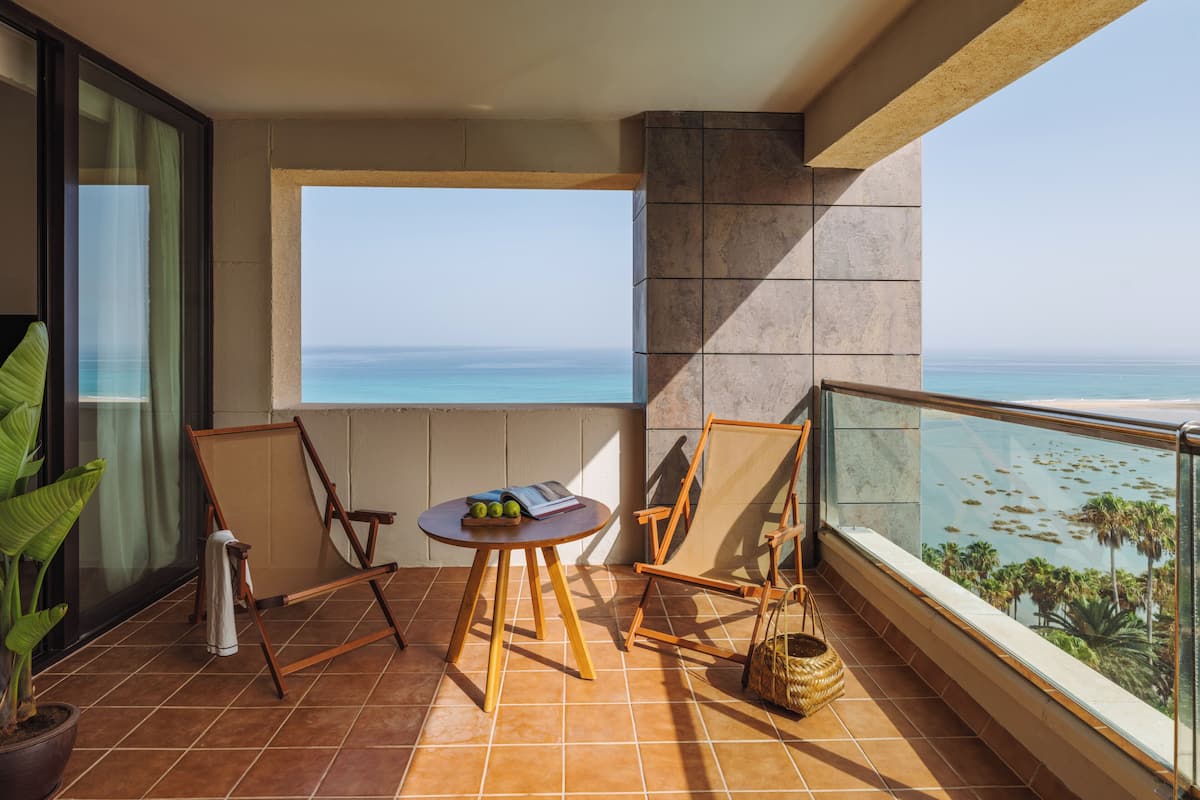 A balcony with a view of the ocean, a table with a book and apples, and a chair.