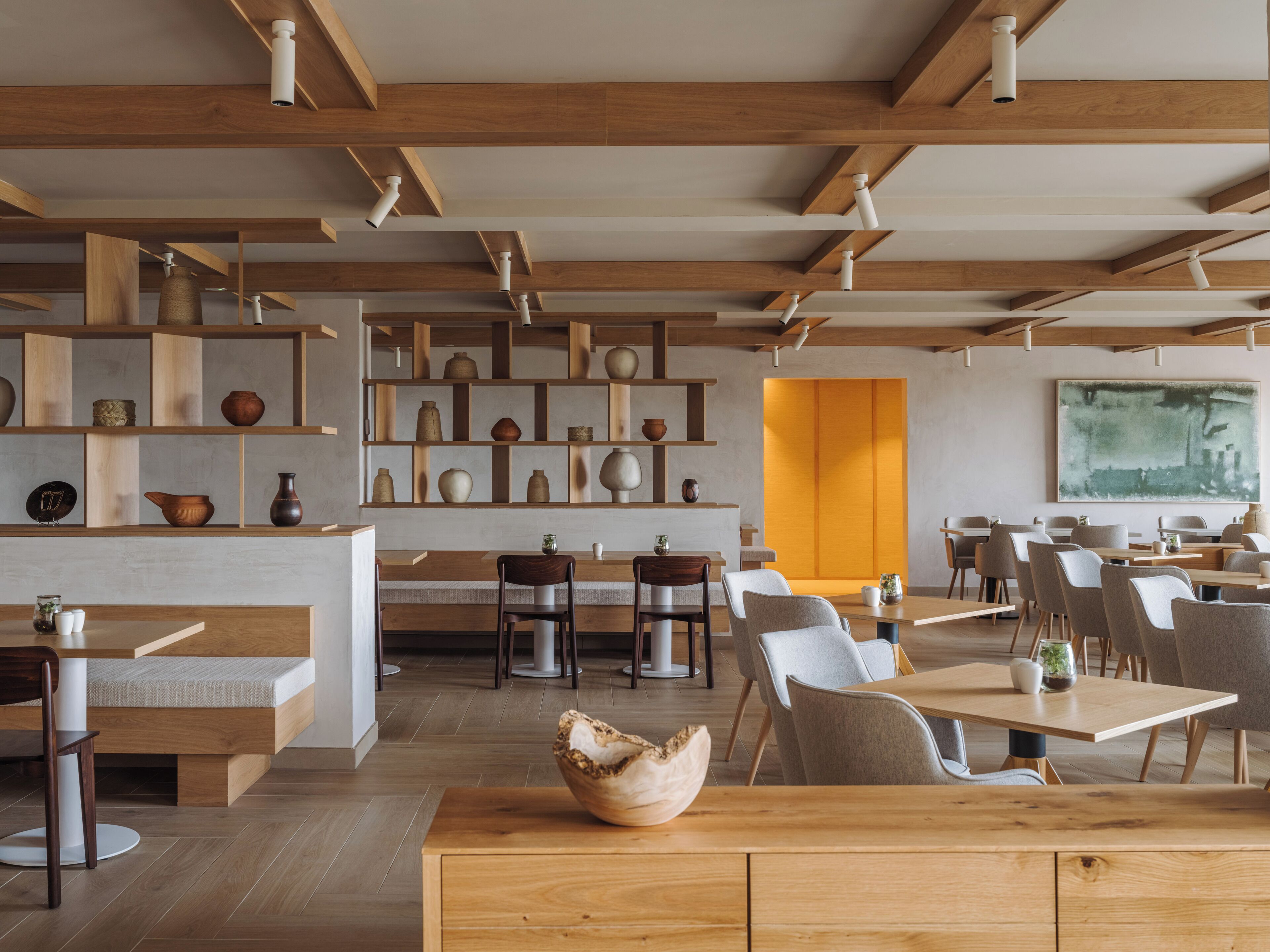 A modern restaurant interior with wooden beams, shelves with pottery, and a bright yellow accent wall.