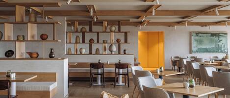 A modern restaurant interior with wooden beams, shelves with pottery, and a bright yellow accent wall.