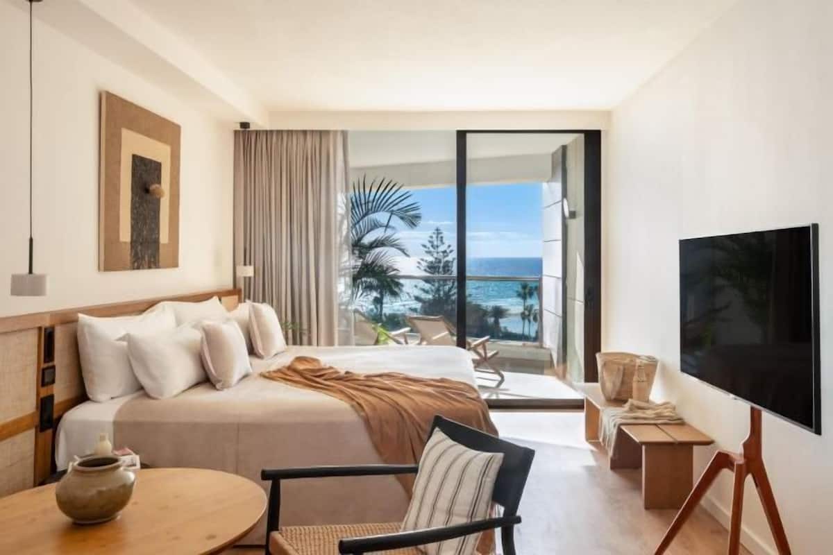 A hotel room with a bed, a TV, a table, and a view of the ocean.