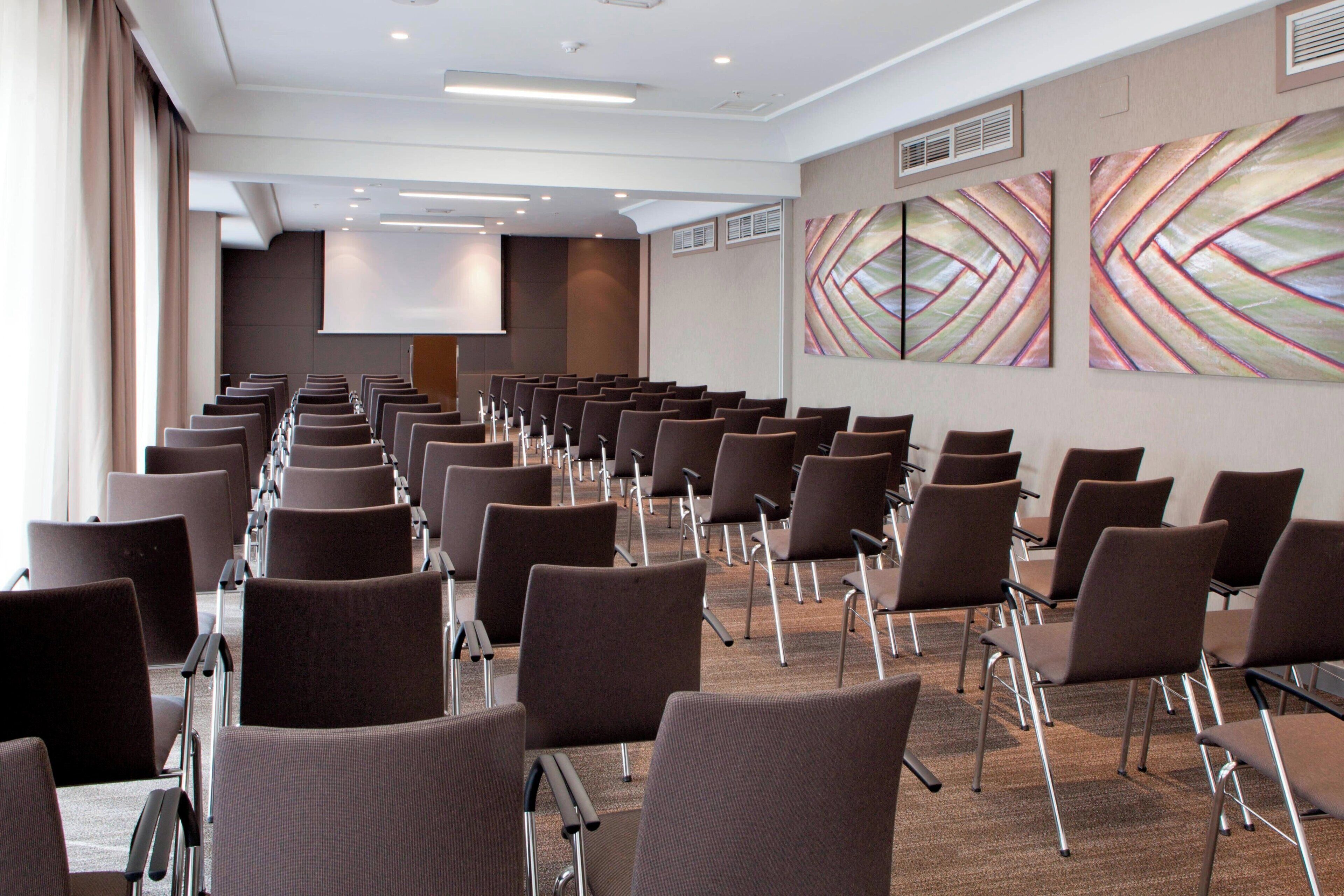 A conference room with rows of chairs, a projector screen, and abstract wall art.