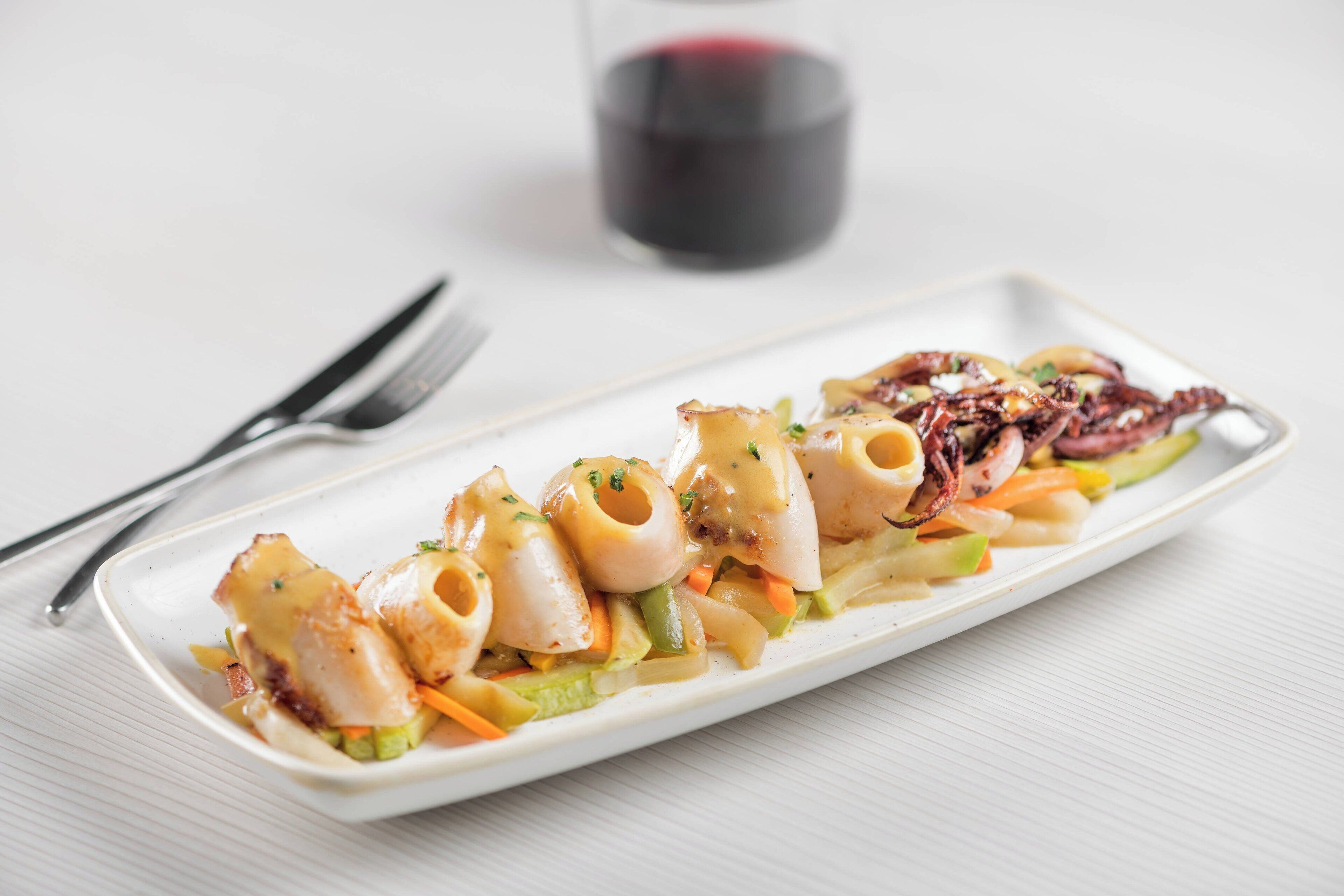 Plato blanco rectangular con pasta y vegetales, acompañado de una copa de vino tinto y cubiertos sobre una superficie blanca.