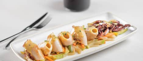 Plato blanco rectangular con pasta y vegetales, acompañado de una copa de vino tinto y cubiertos sobre una superficie blanca.