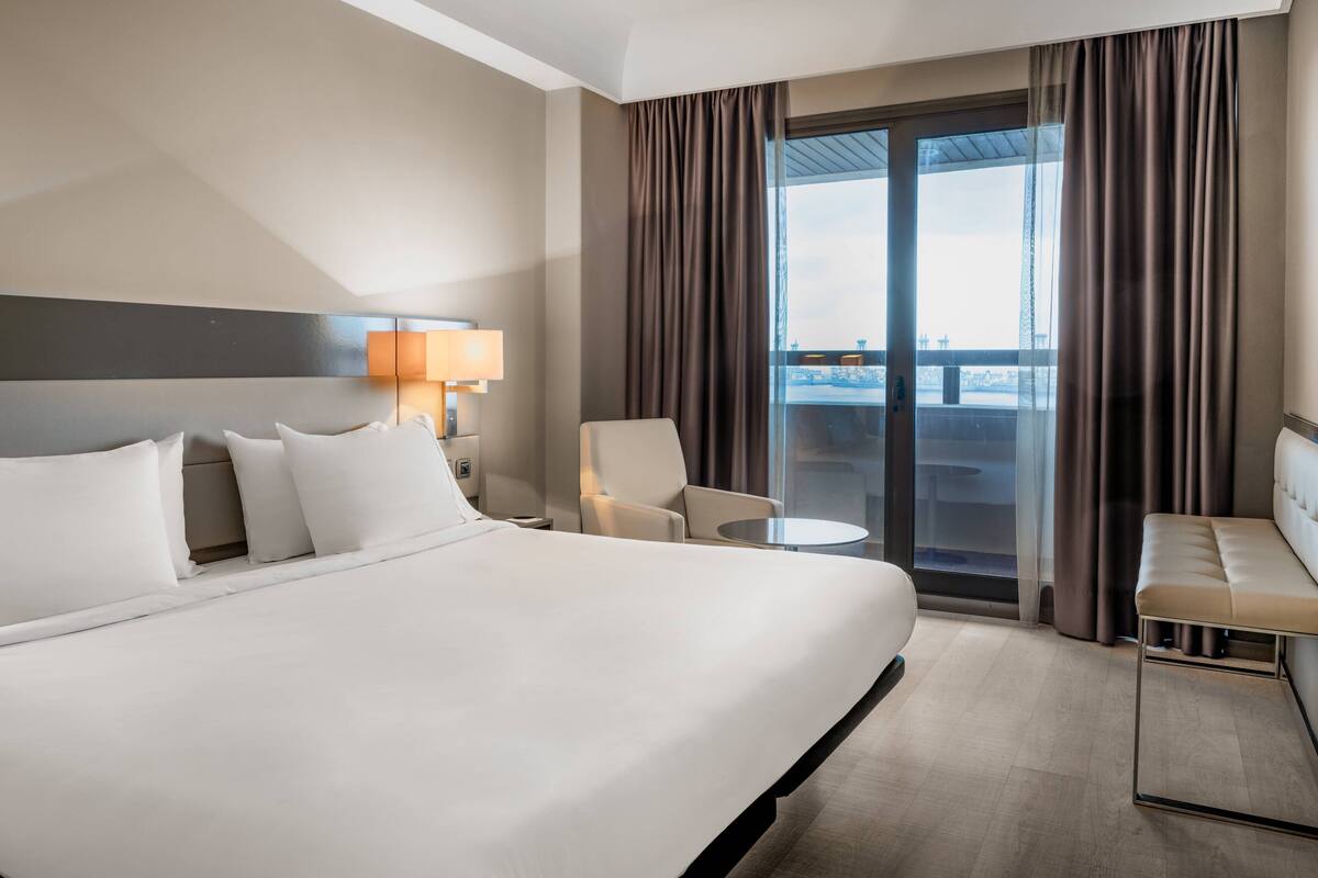 Une chambre d’hôtel avec un grand lit, un bureau, une chaise et une vue sur la ville grâce à une grande fenêtre.