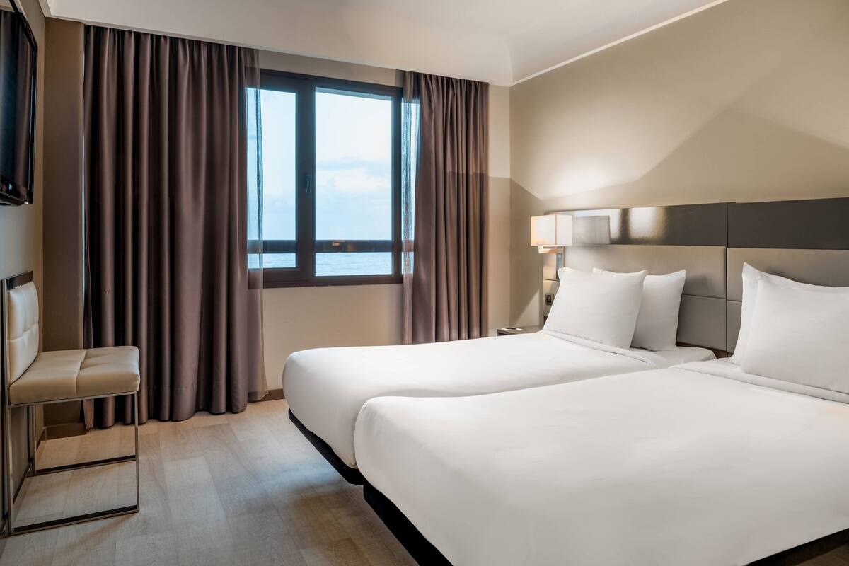 Une chambre d’hôtel avec deux lits, un bureau, une chaise et une grande fenêtre avec des rideaux.