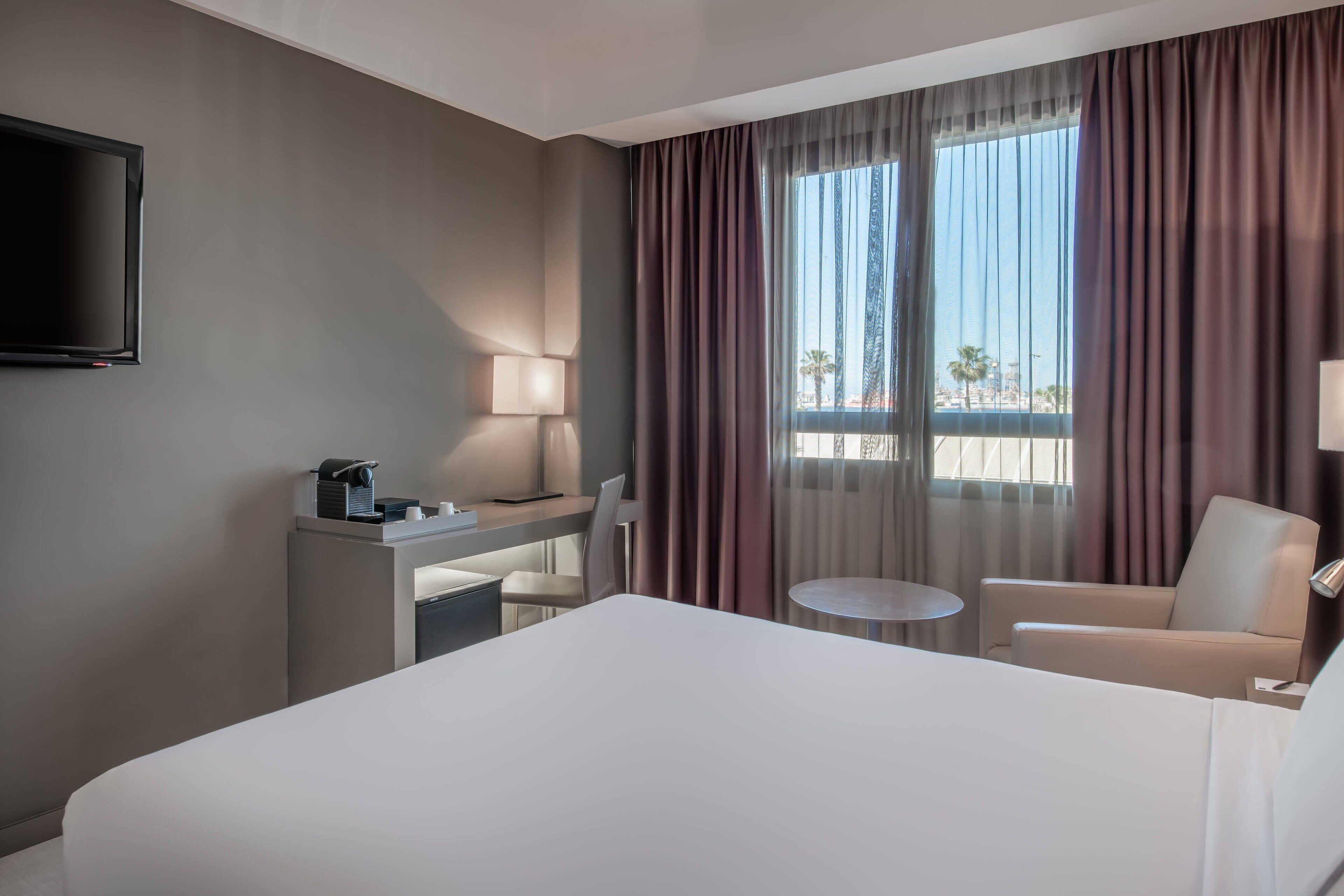 Una habitación de hotel con una cama, un escritorio con lámpara, una silla, una mesita y un televisor montado en la pared.