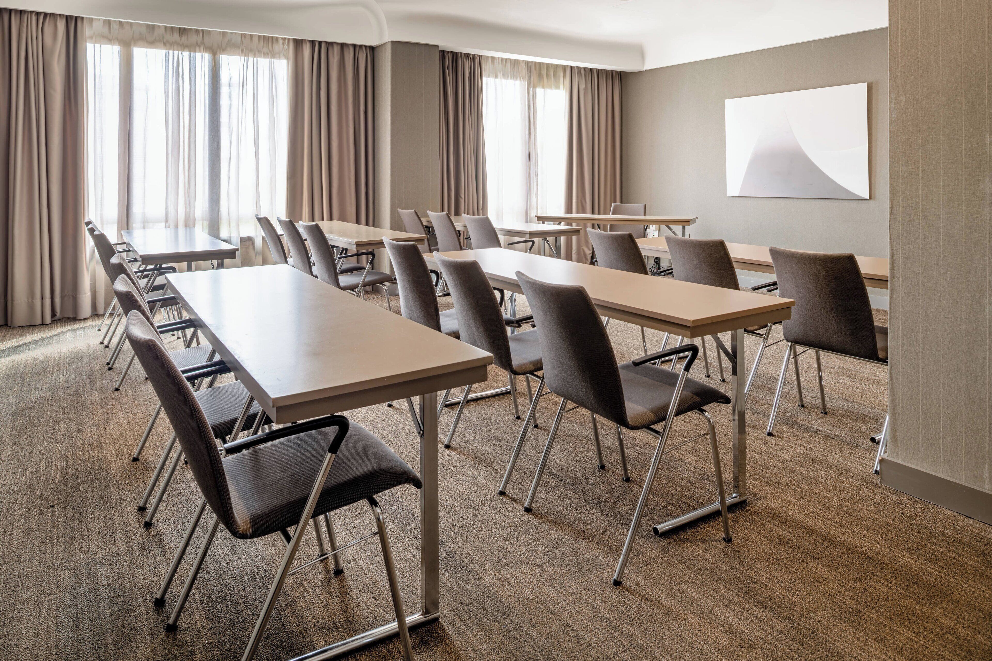 A conference room with multiple tables and chairs, large windows with curtains, and a plain wall with a single artwork.