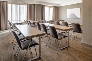 A conference room with multiple tables and chairs, large windows with curtains, and a plain wall with a single artwork.