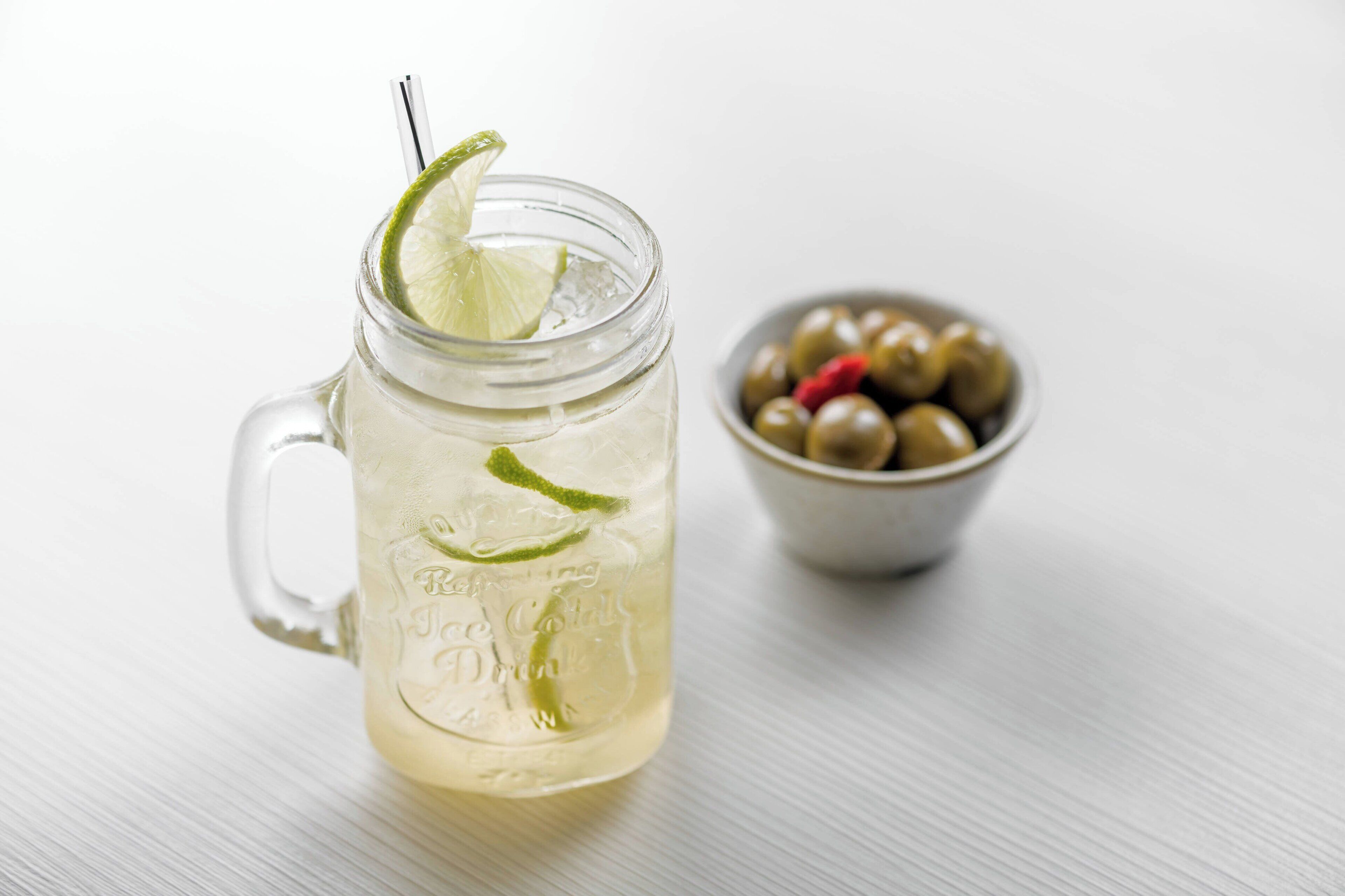 A glass mug with a lime slice and straw, labeled "Refreshing Juice Cocktail," next to a bowl of olives.