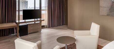 A modern hotel room with a large window, a flat-screen TV, a desk, two white armchairs, a small round table, and a brown rug.
