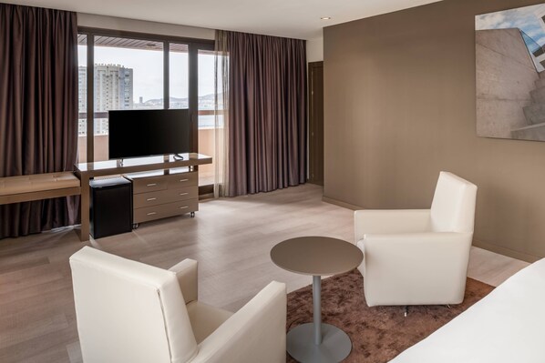 Une chambre d’hôtel moderne dotée d’une grande fenêtre, d’un téléviseur à écran plat, d’un bureau, de deux fauteuils blancs, d’une petite table ronde et d’un tapis marron.
