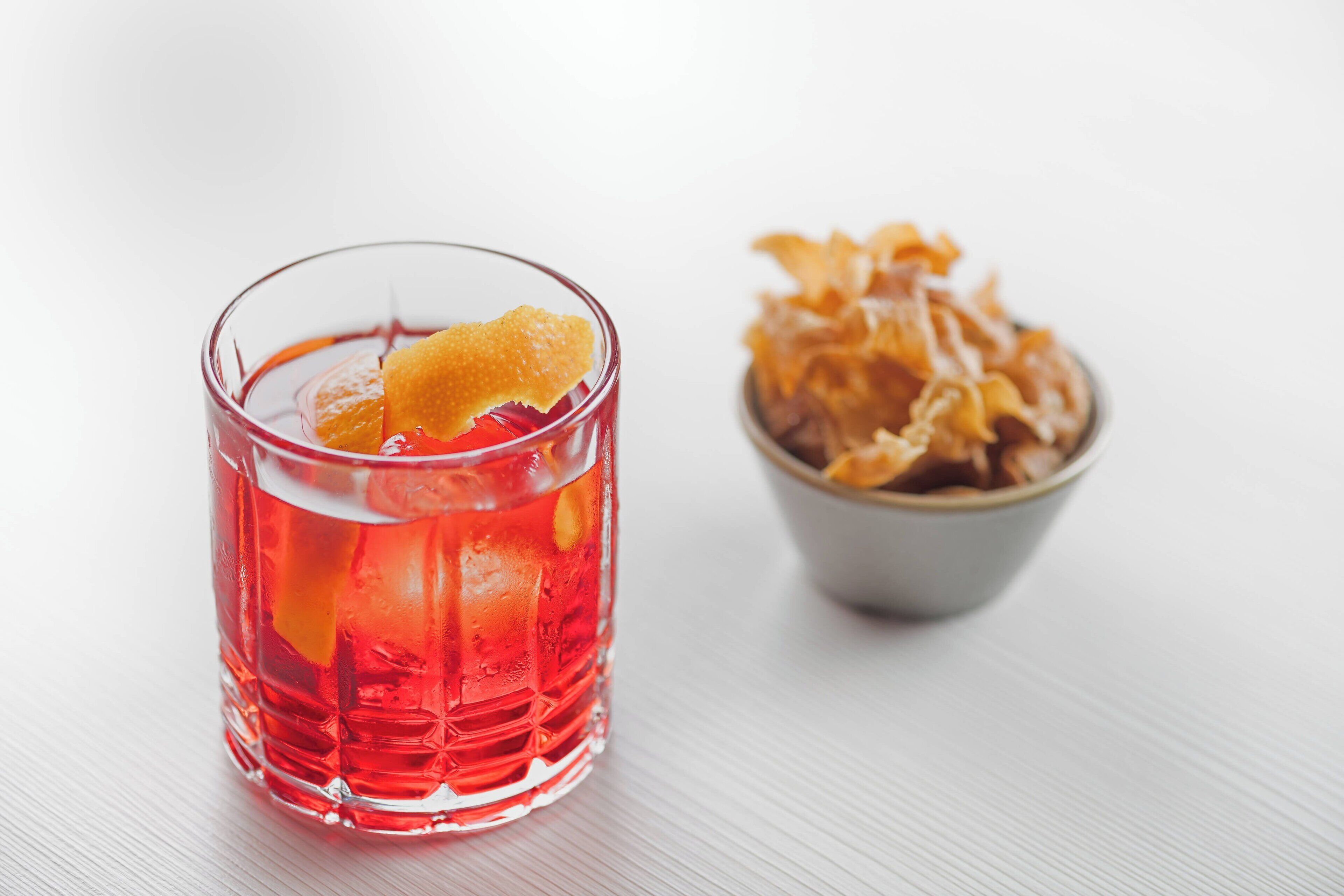 Un verre transparent contenant une boisson rouge et décoré d’une tranche d’agrumes, accompagné d’un bol de fruits secs.