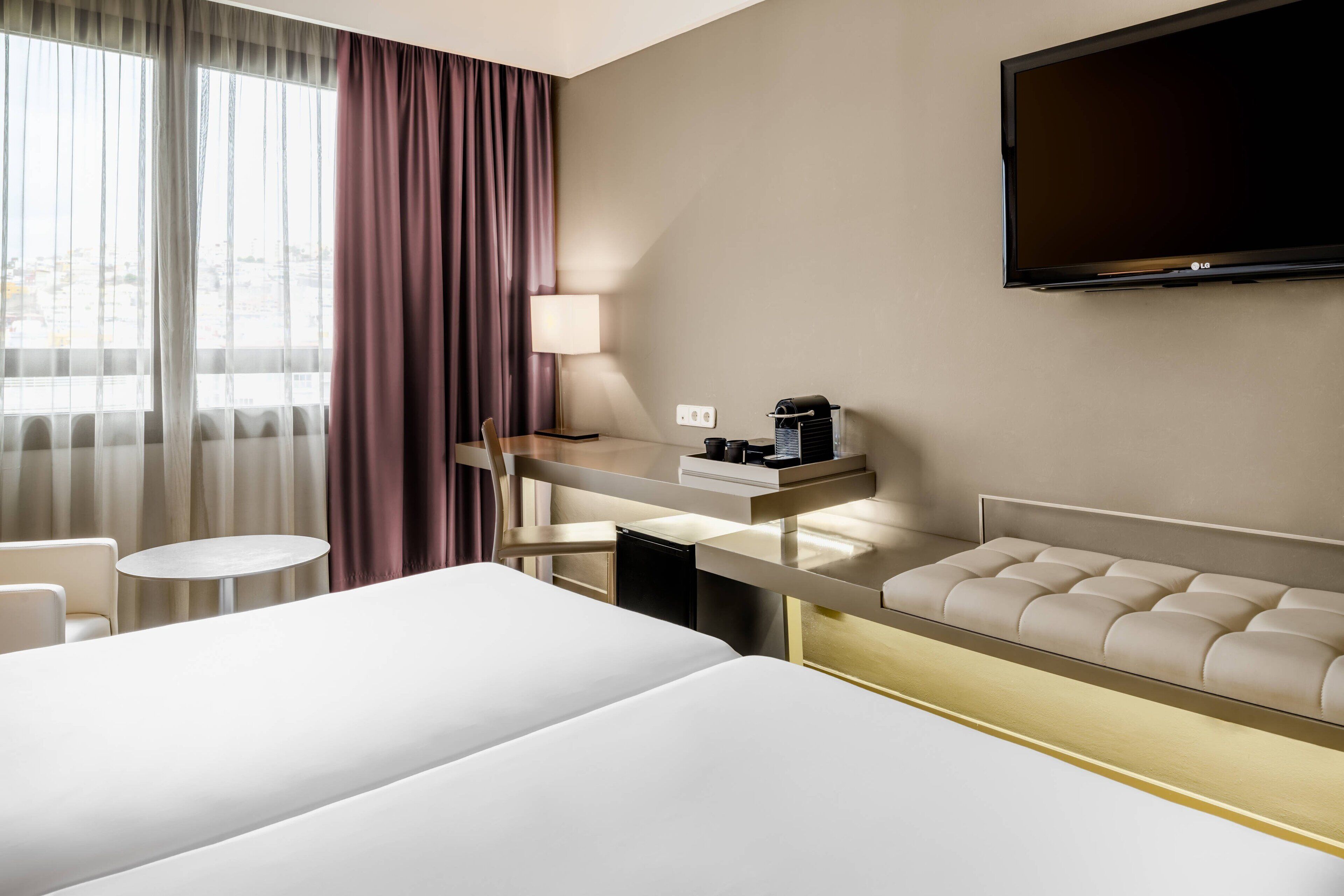 A hotel room with a large bed, a desk with a lamp, a television, and a view of the city through the window.