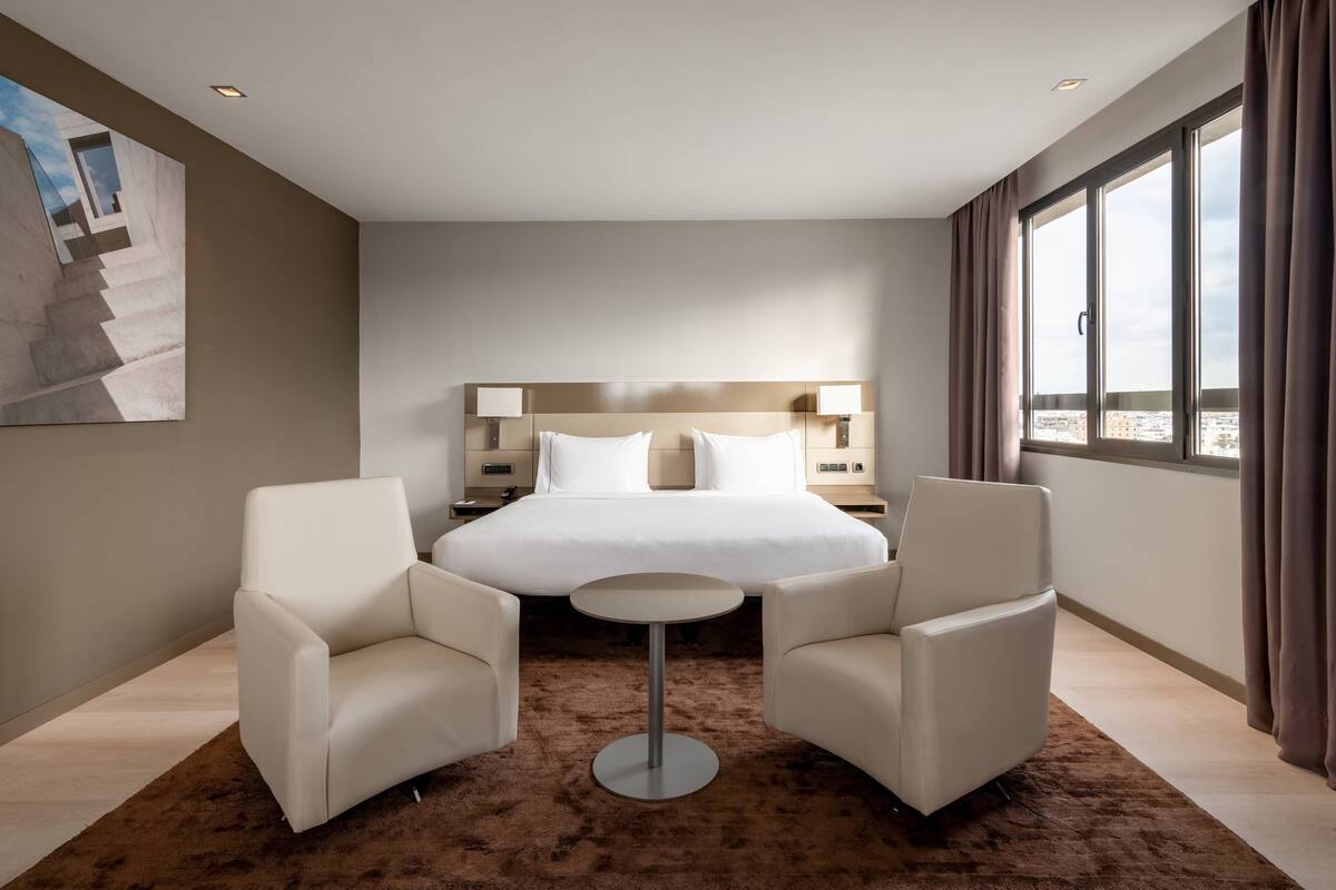 Ein modernes Hotelzimmer mit einem großen Bett, zwei Sessel, einem kleinen runden Tisch und Blick auf die Stadt durch große Fenster.