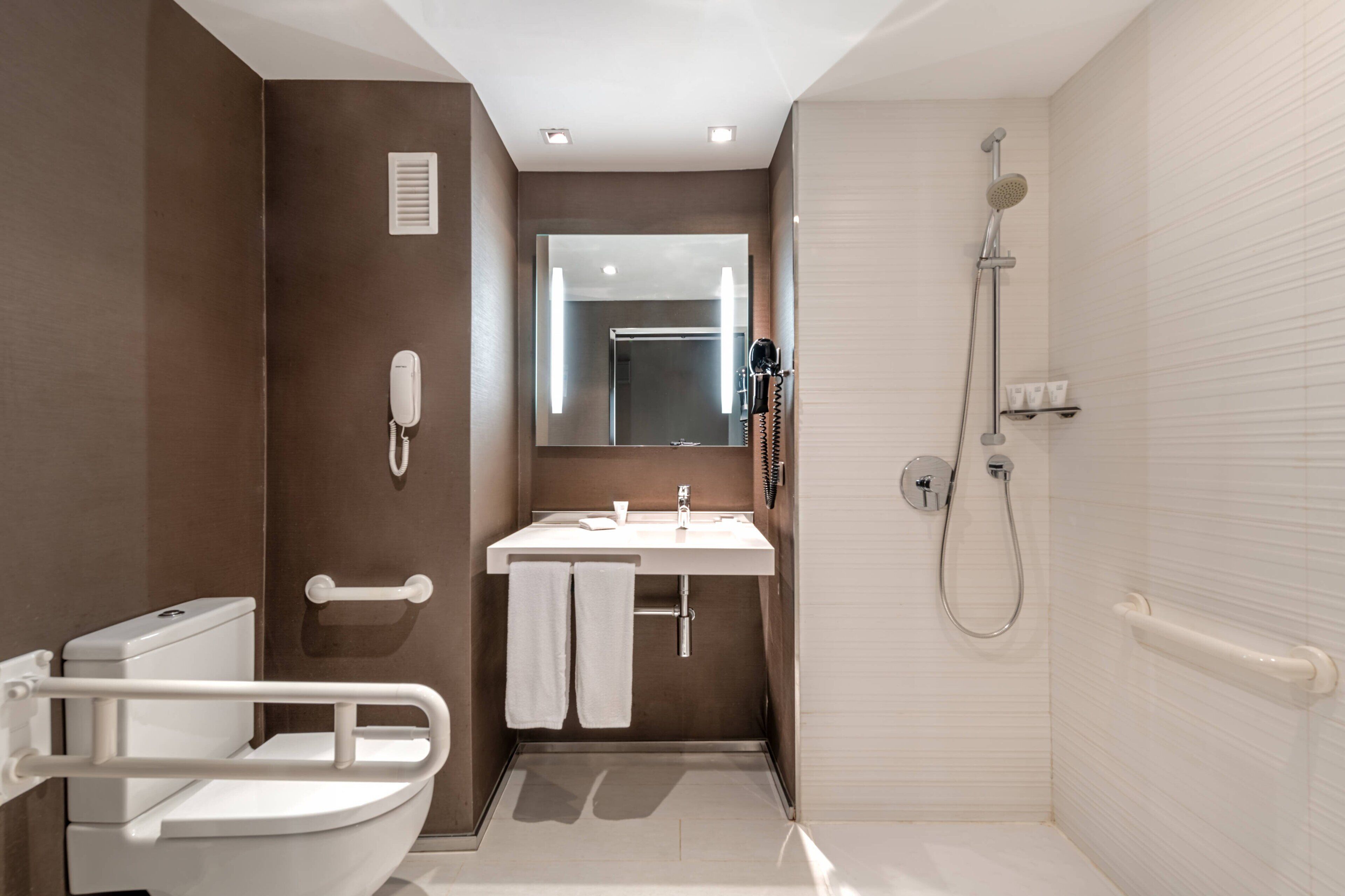 A modern bathroom with a toilet, sink, and shower. It includes a handrail and a mirror.