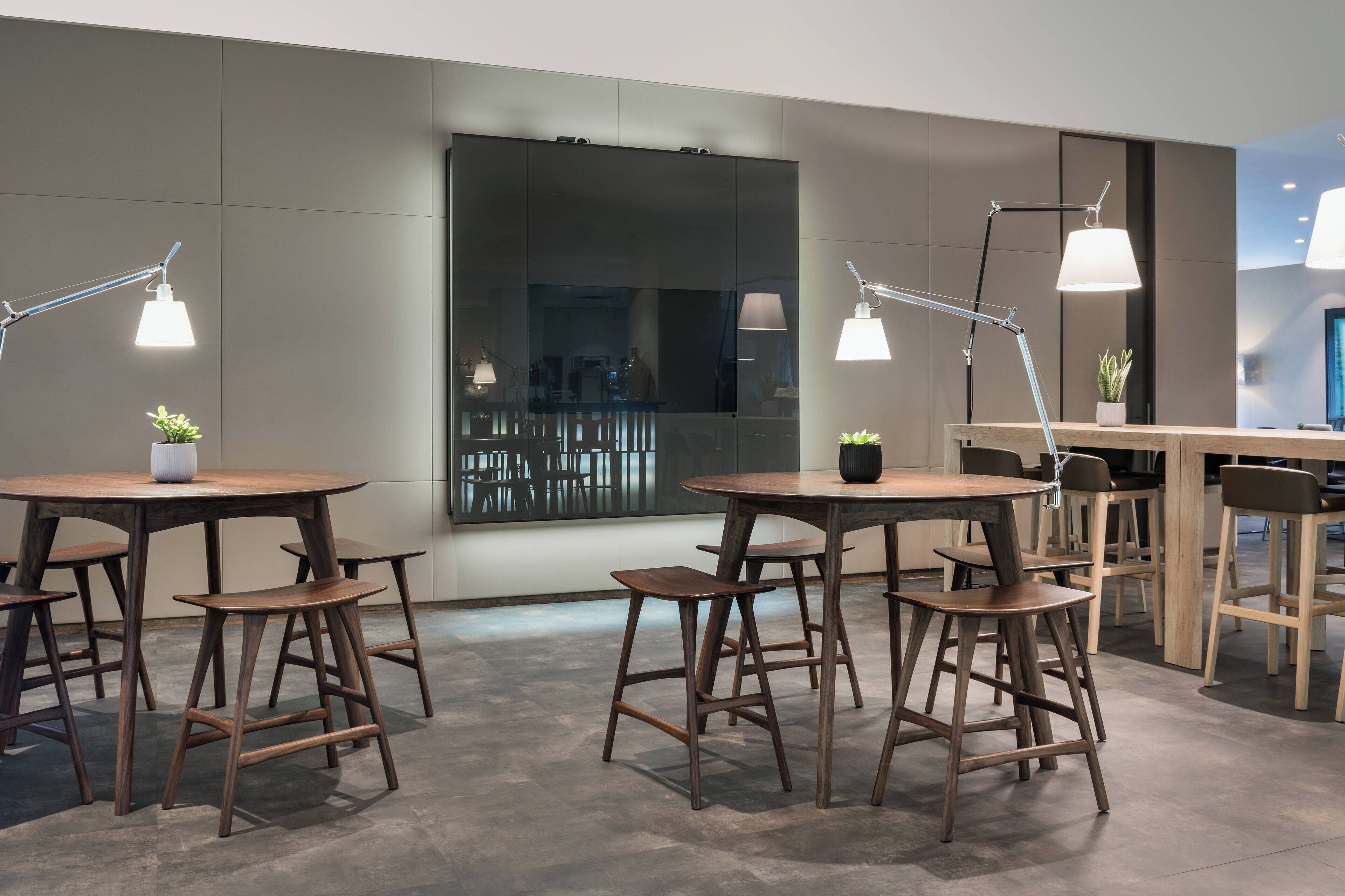 A modern interior with round wooden tables and matching stools, two pendant lights, and a large window with a view of outdoor seating.