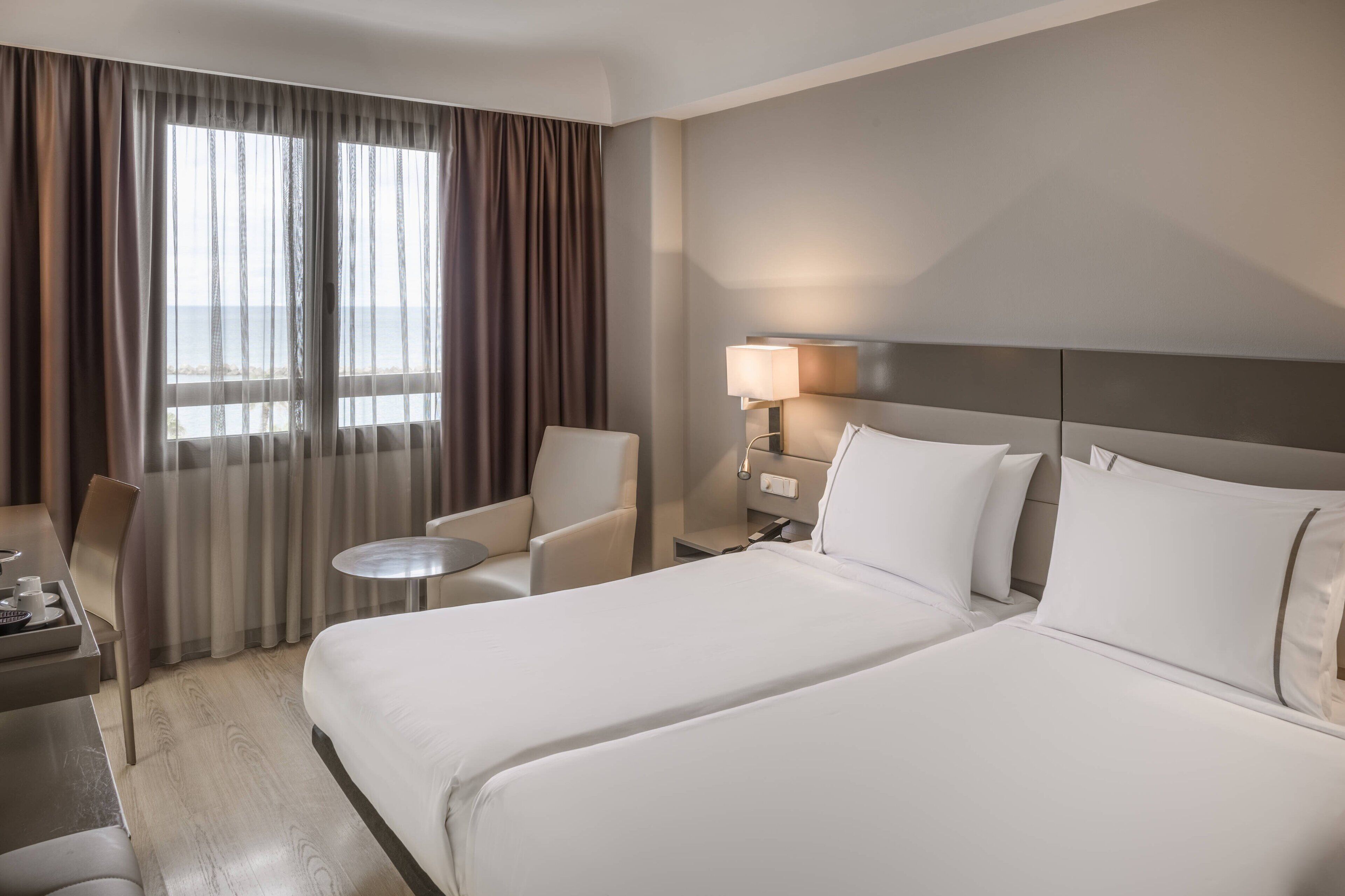 A hotel room with a large bed, a desk with a chair, a small round table, and a lamp.