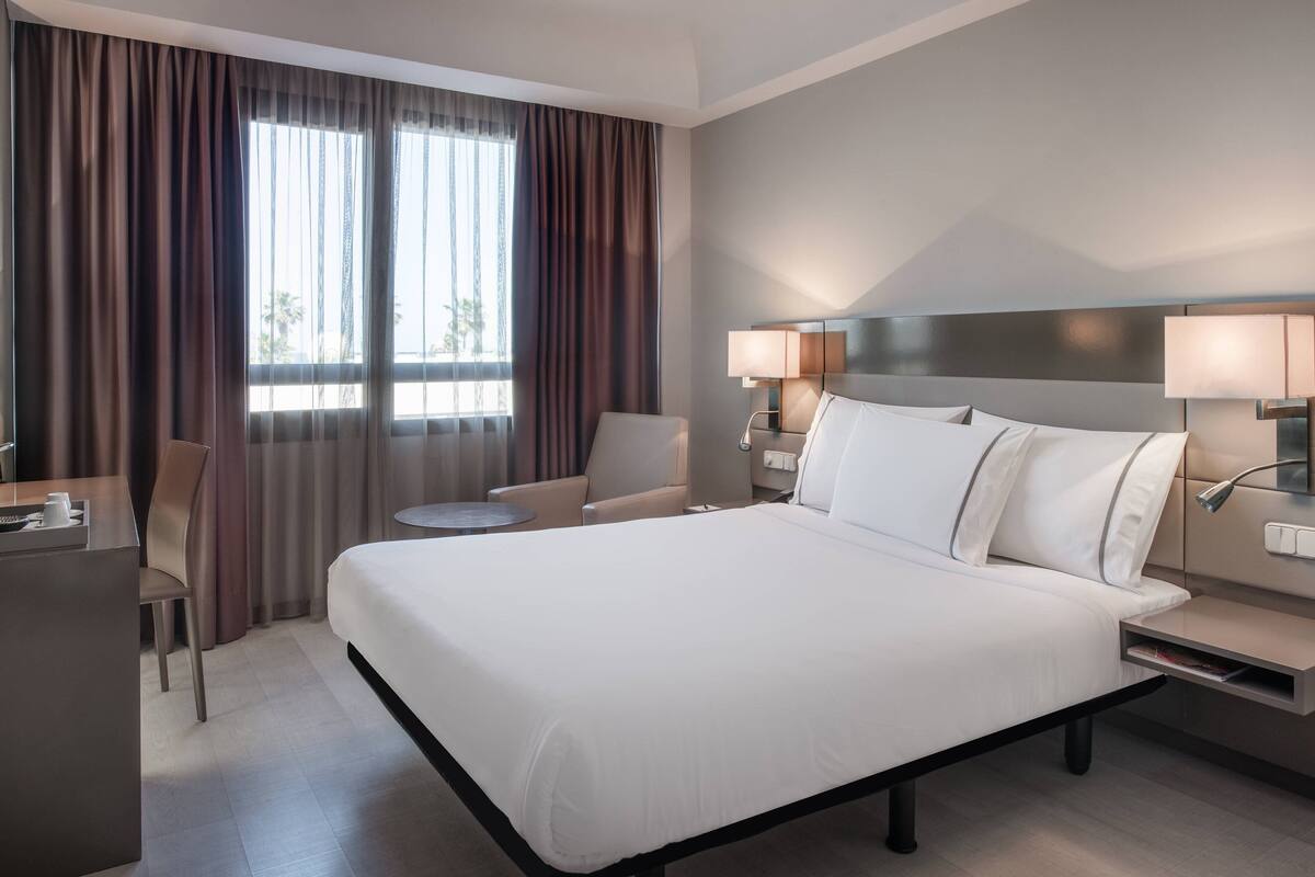 Une chambre d’hôtel avec un grand lit, deux tables de chevet avec des lampes, un bureau avec une chaise et une fenêtre avec des rideaux.