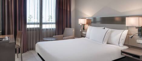 A hotel room with a large bed, two bedside tables with lamps, a desk with a chair, and a window with curtains.