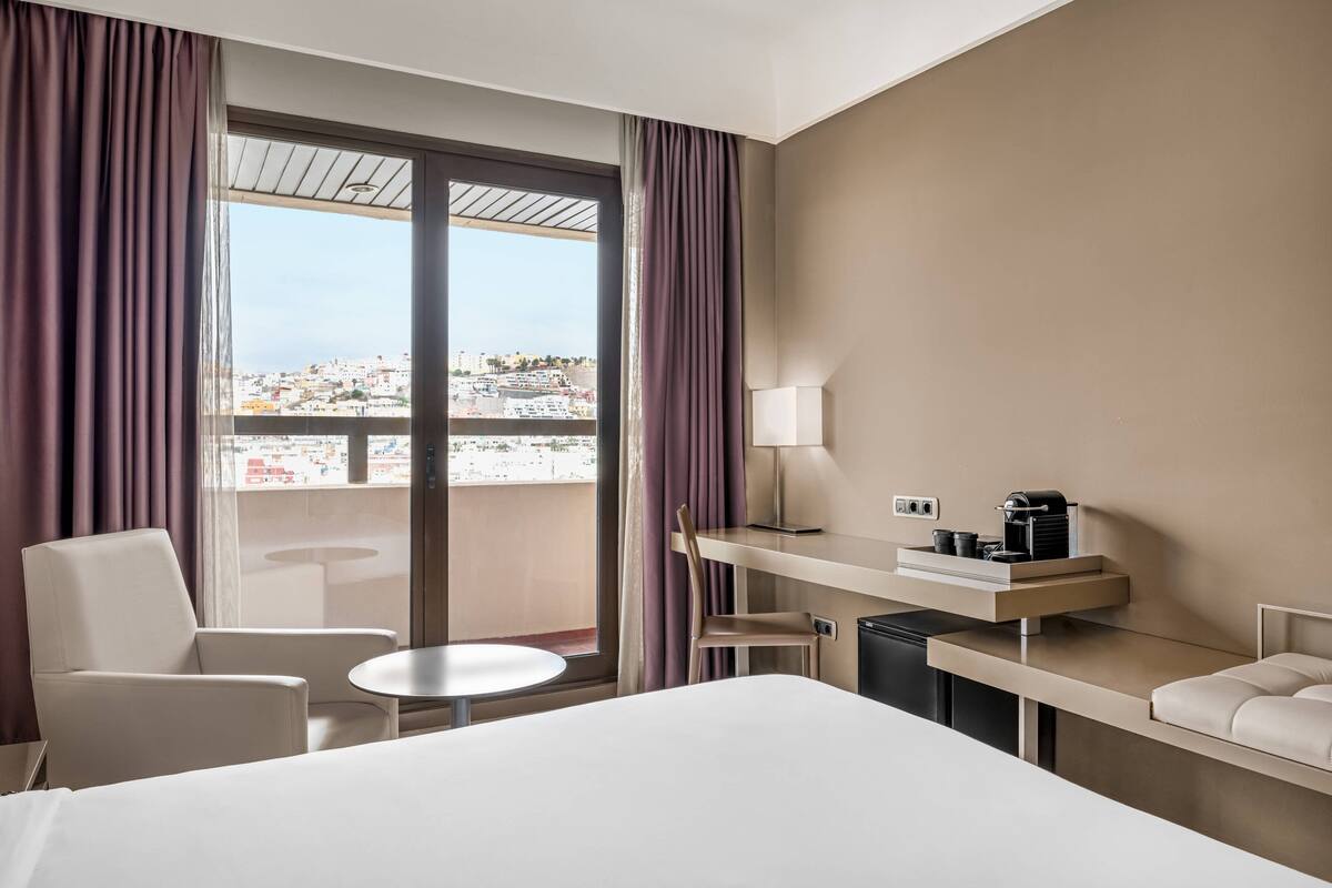 Une chambre d’hôtel comprenant un lit, un bureau avec une machine à café, une chaise, une petite table et une vue sur le paysage urbain grâce à de grandes fenêtres.